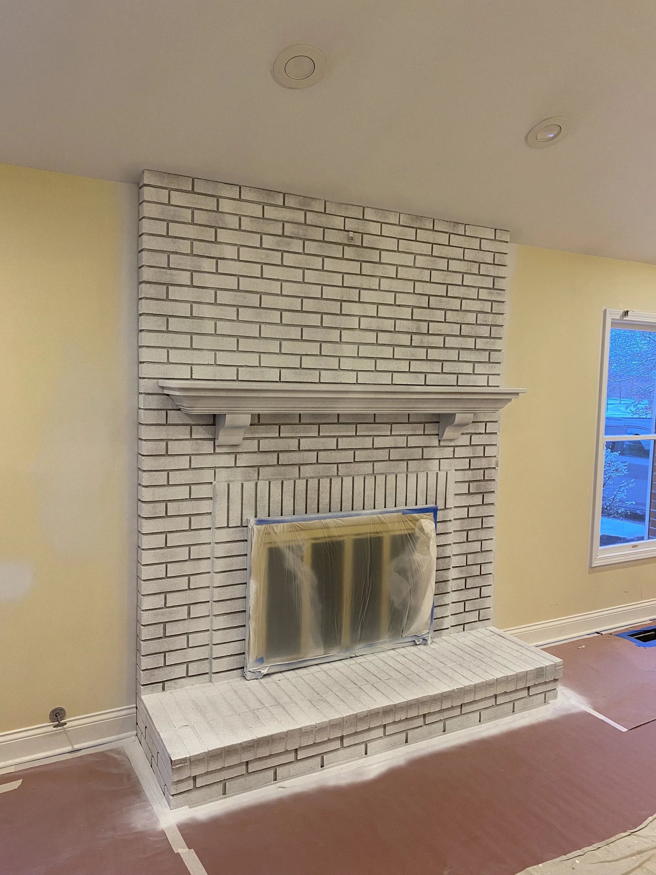 A brick fireplace with a mantel, partially covered in plastic, in a room with yellow walls and a window on the right side.