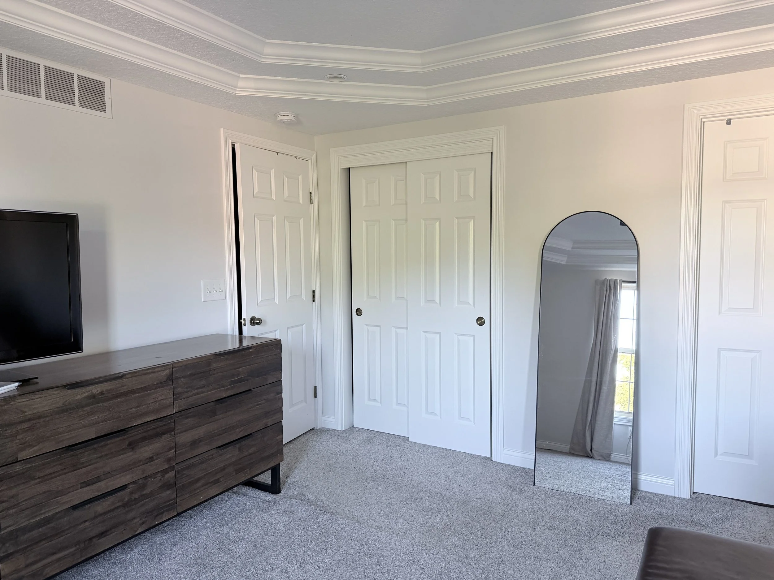 Living room corner with white walls, a ceiling with crown molding, a closed double closet door, a black TV, a wooden dresser, a mirror, a window with curtains, and a gray carpeted floor.