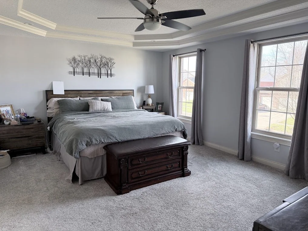 A bedroom with a large bed, two nightstands, and two windows letting in natural light. The room has a ceiling fan and a grey and white color scheme.