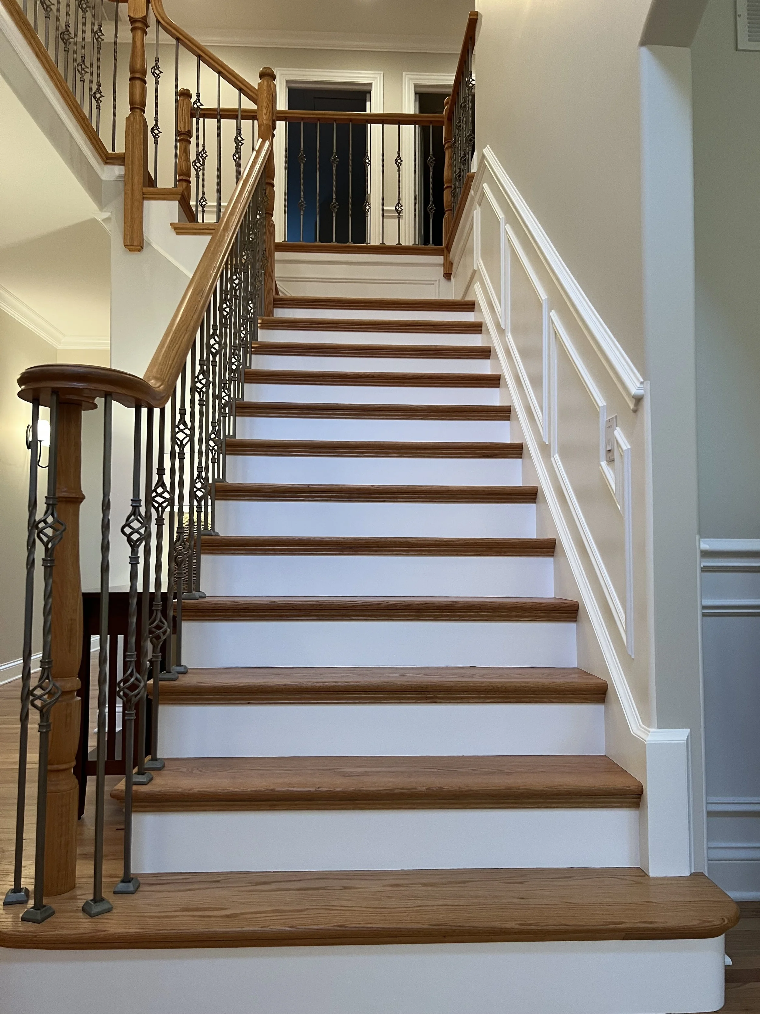 Wooden staircase with white risers, wooden handrail, and decorative black metal balusters inside a home, leading to a small landing with a black door.