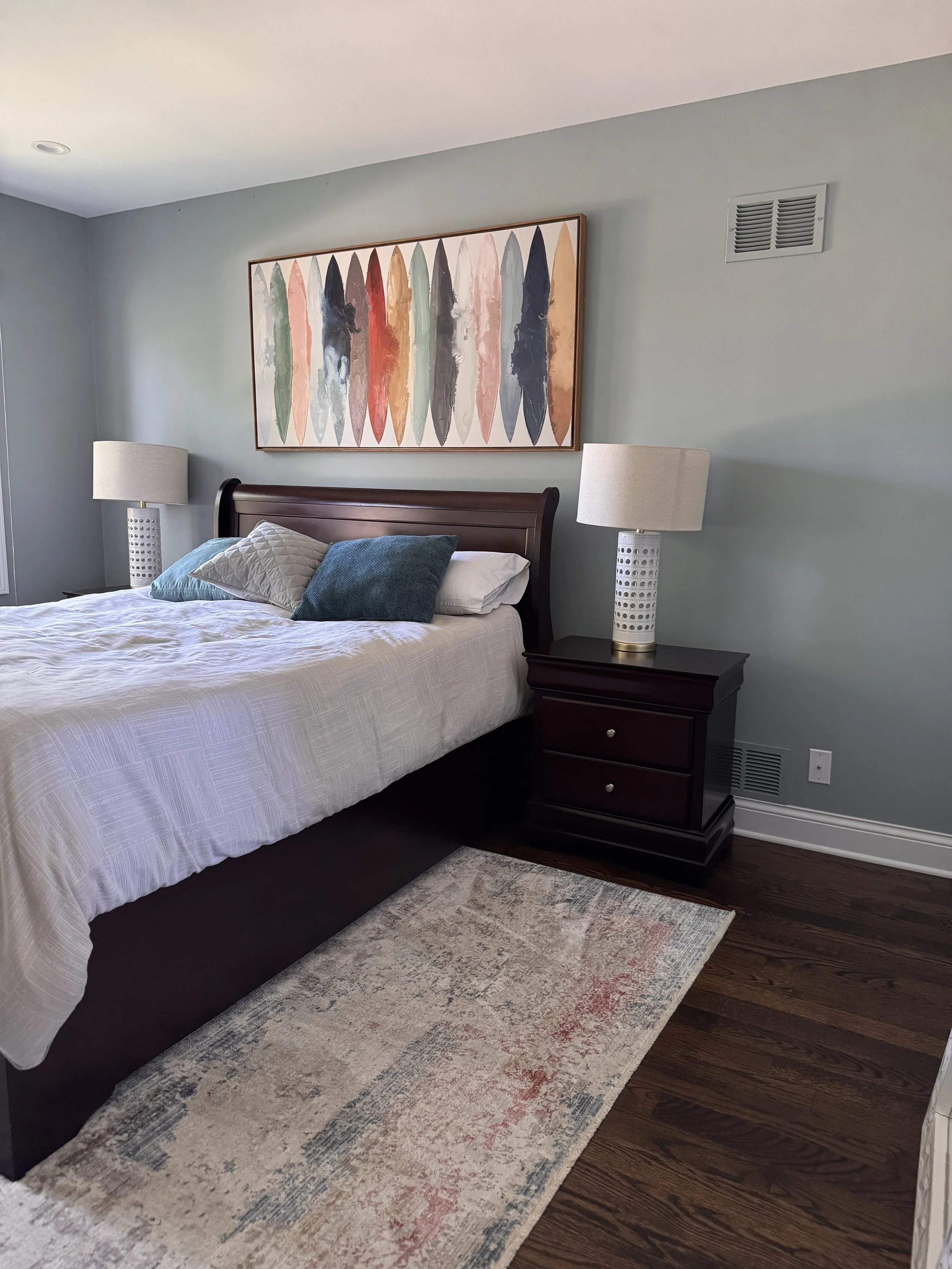 A bedroom with a wooden bedframe, white bedspread, and decorative pillows. Nightstands with white lamps sit on either side of the bed. Above the bed hangs a large abstract painting with colorful elongated shapes. A rug with faded multicolored patterns lies on dark hardwood floor. The wall is painted light green and features a ventilation grate.