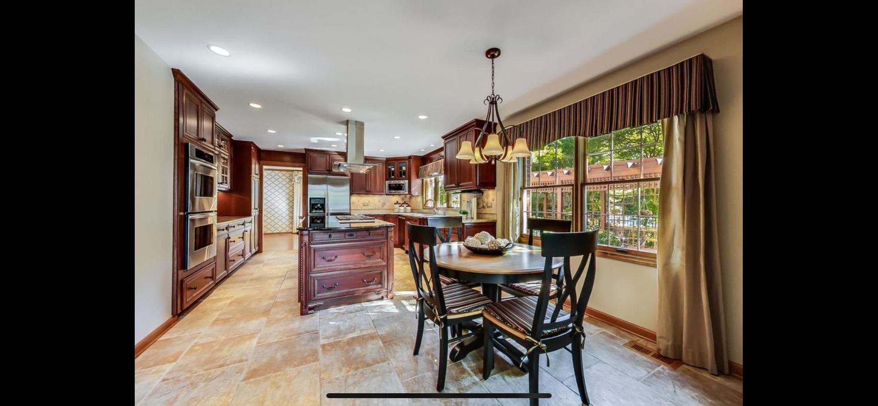 A spacious kitchen with wooden cabinets, a dining table with four chairs, large windows with striped valances, and a chandelier light fixture.
