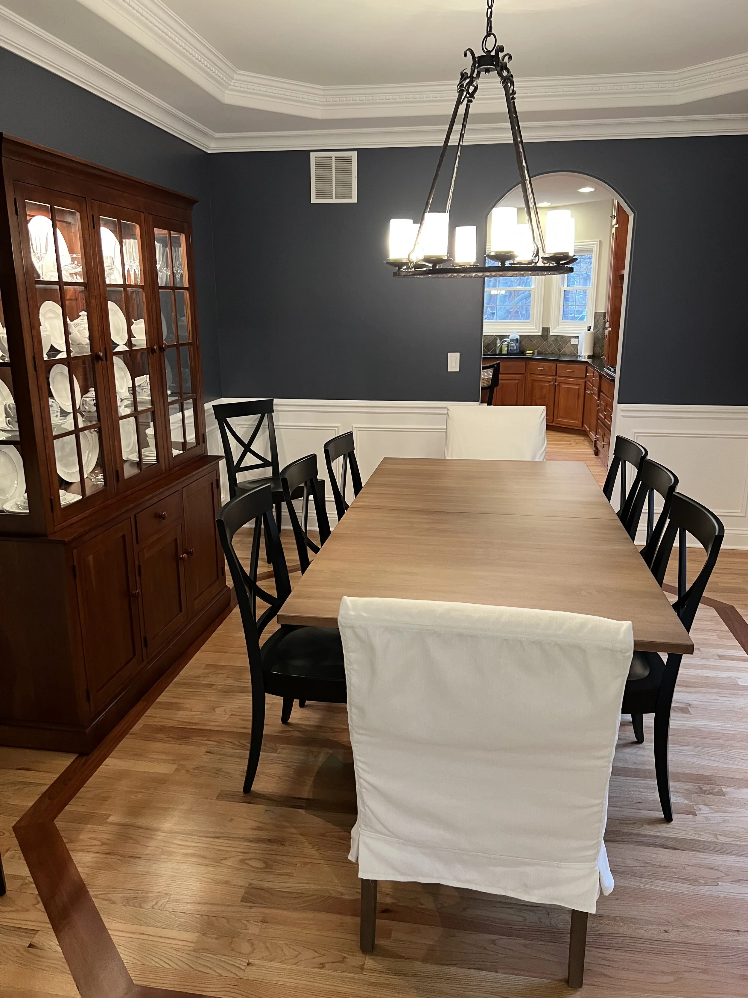 A dining room with a wooden table and six chairs, four of which are black and two with white slipcovers. A wooden china cabinet filled with white dishware is on the left. The back wall is painted dark blue with white trim, and there is an entry to the kitchen with wooden cabinets and a window showing trees outside. A chandelier hangs above the table.