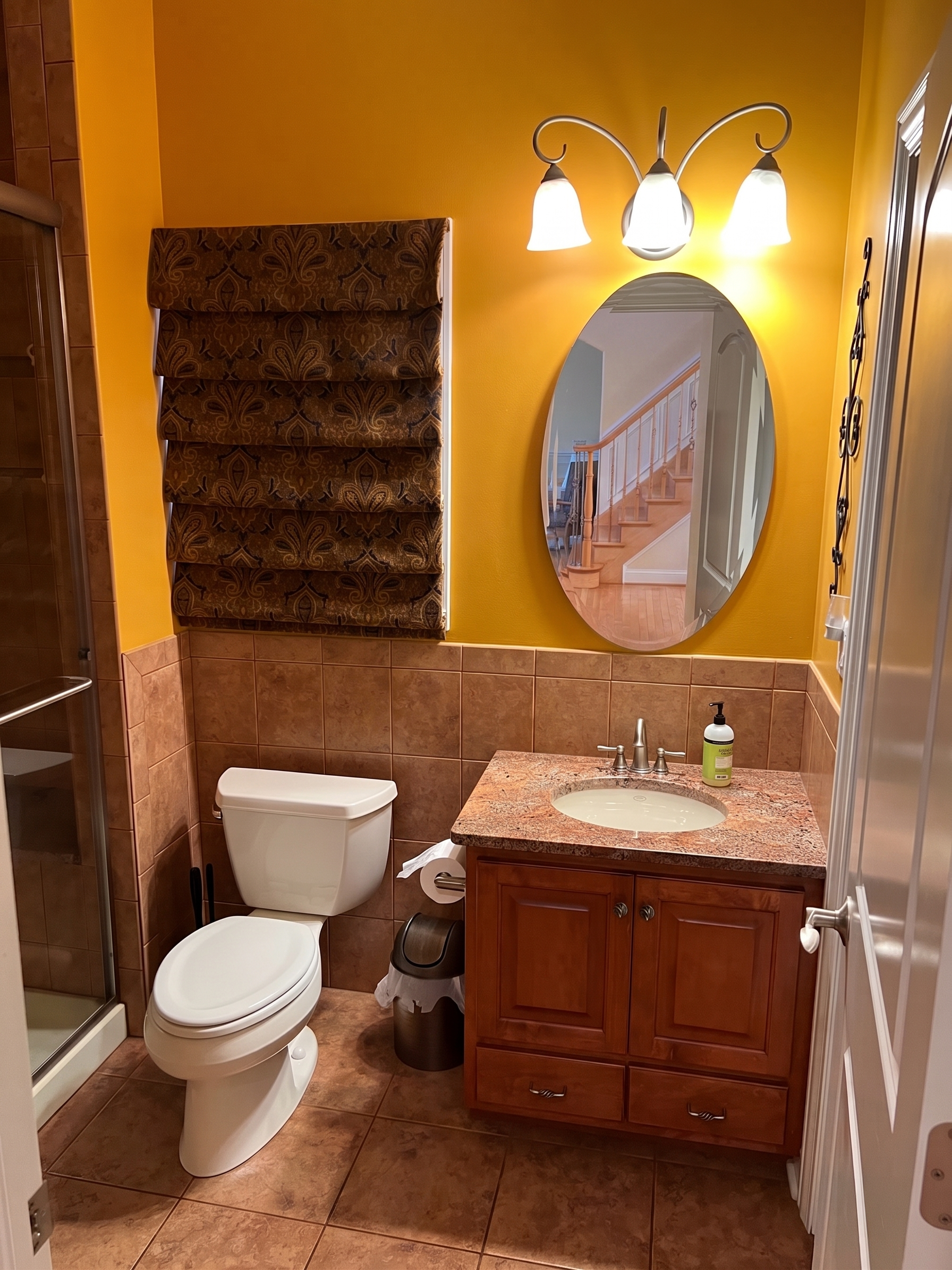Bathroom with yellow walls, a toilet, a vanity with a granite countertop and an oval mirror, and light fixtures above the mirror. There are tiled floors and a partially visible shower.