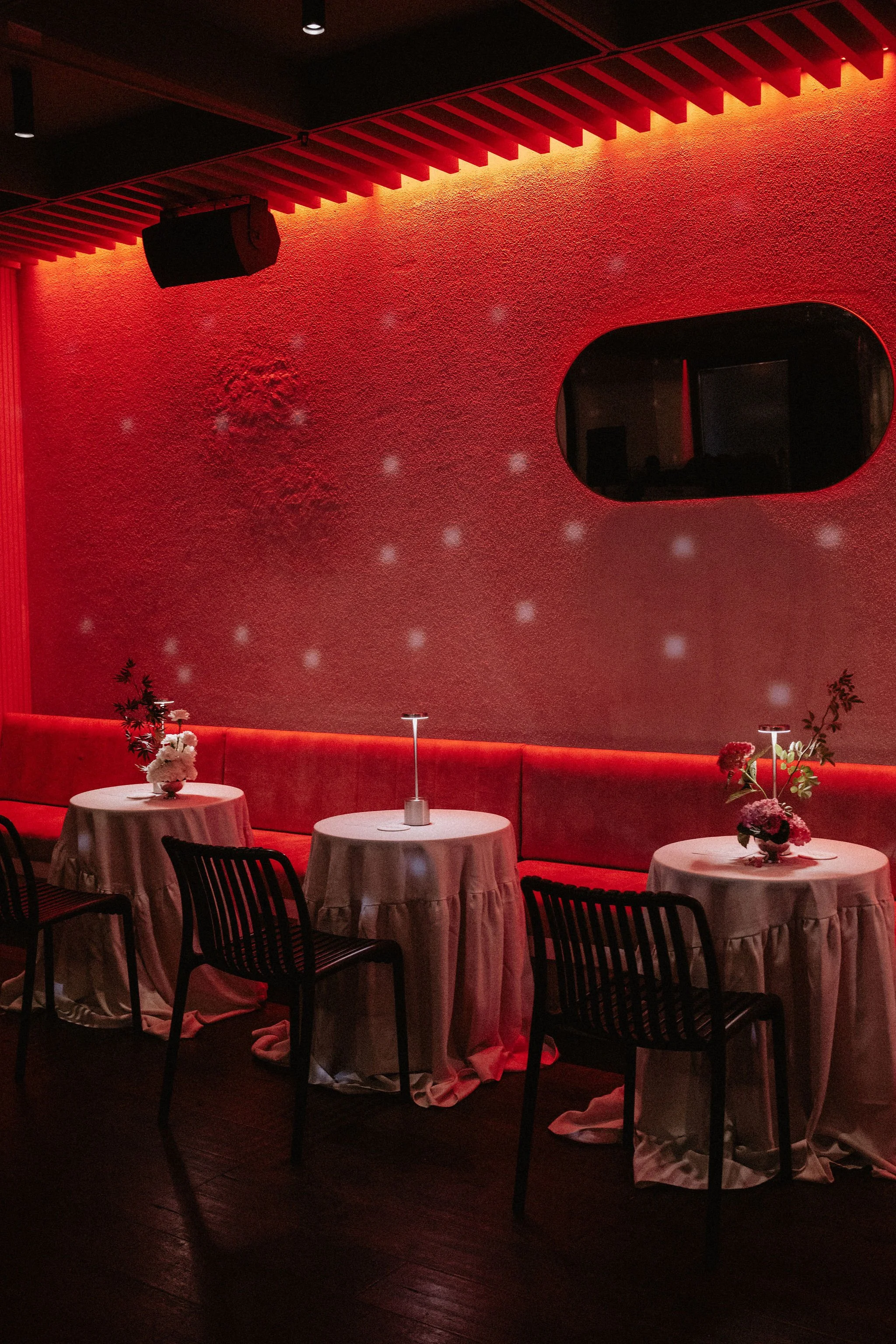 A dimly lit restaurant with small round tables draped in white tablecloths and decorated with floral centerpieces, set against a red textured wall illuminated by a red LED light, with black chairs around the tables and a large oval window on the wall.