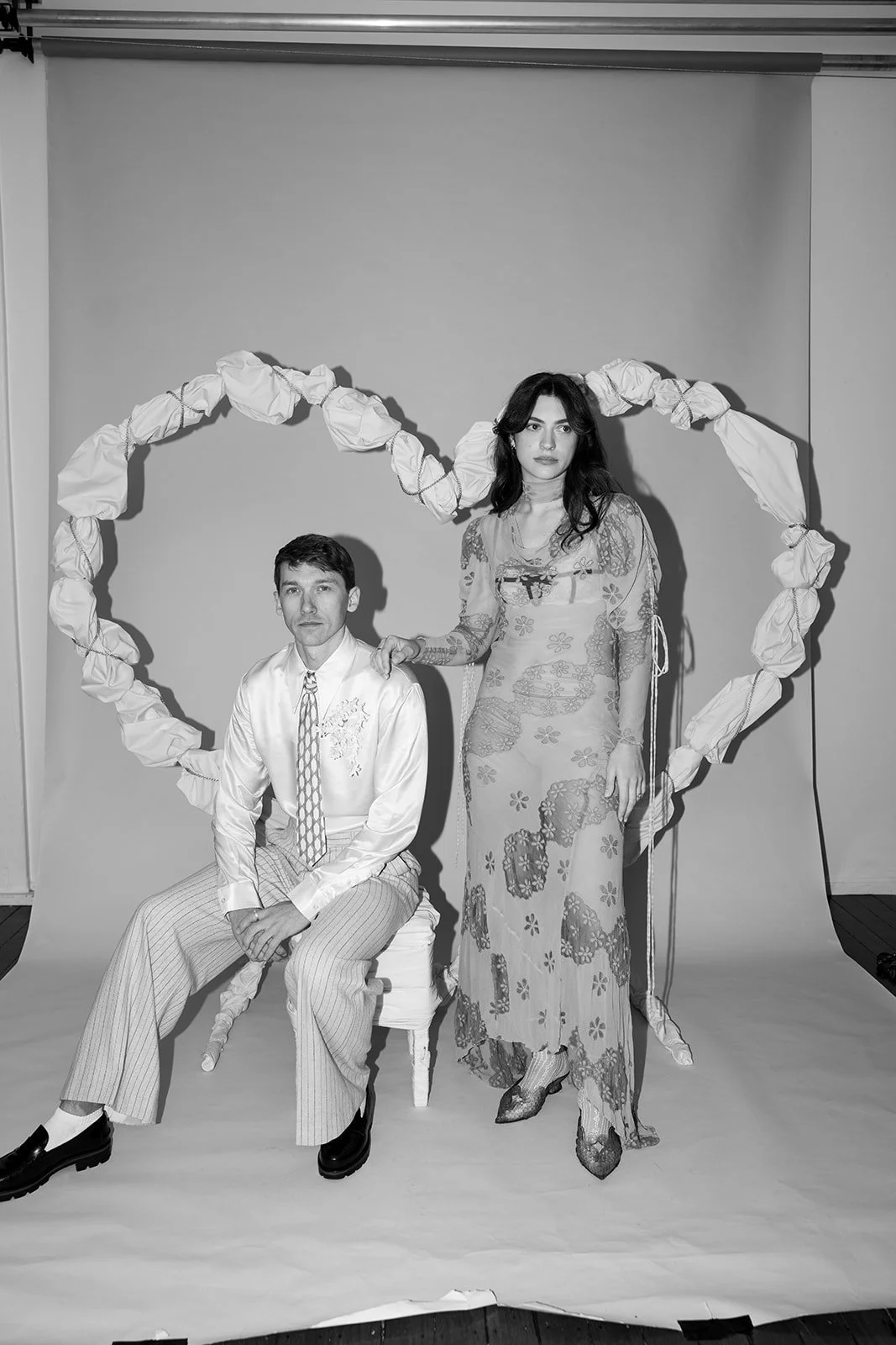 Black and white photo of a man sitting on a chair and a woman standing next to him, with a large heart shape made of fabric behind them.