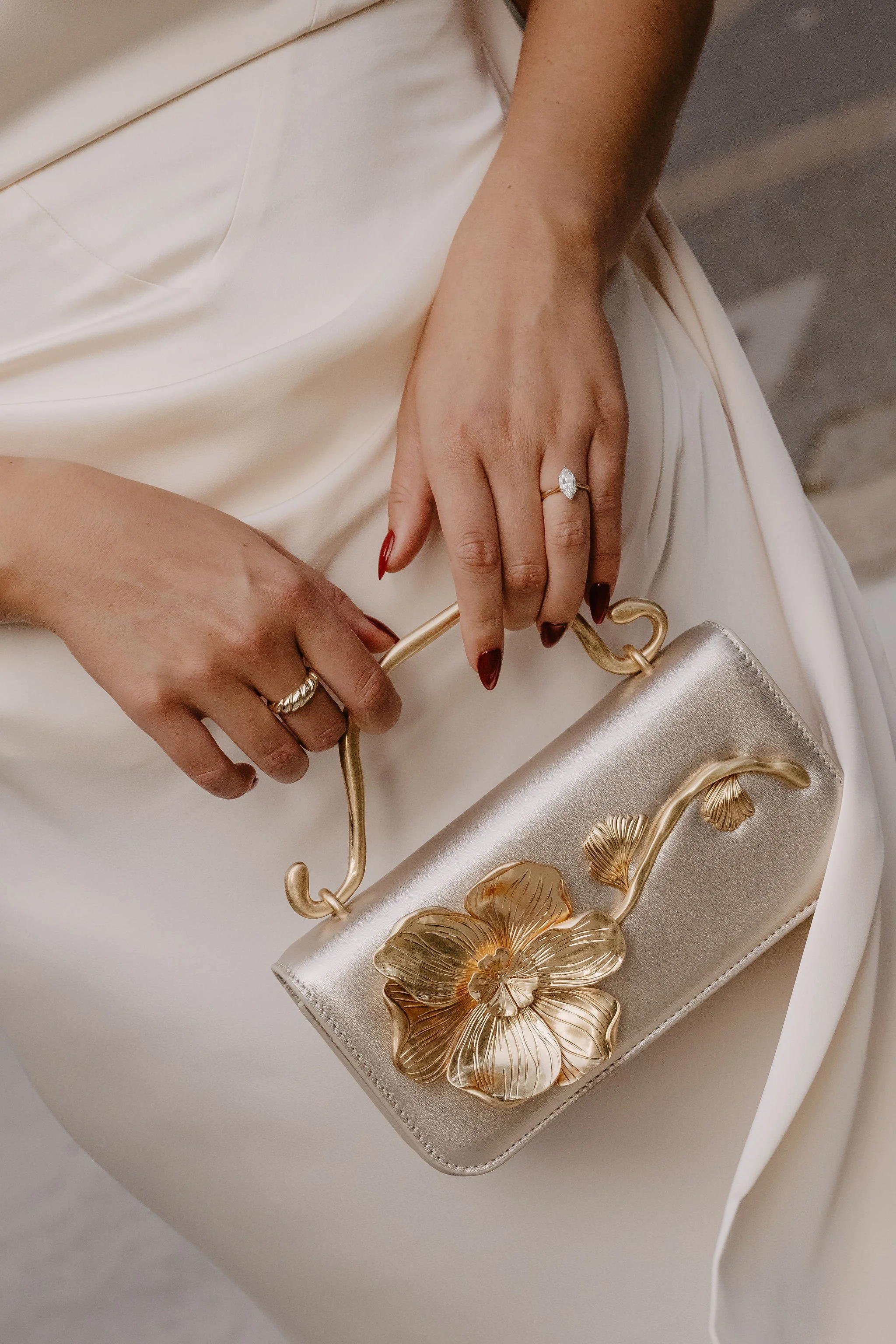 Close-up of a woman's hand with dark red nails, wearing a diamond ring, holding a gold-colored handbag with metallic flower embellishments, against a white satin dress.