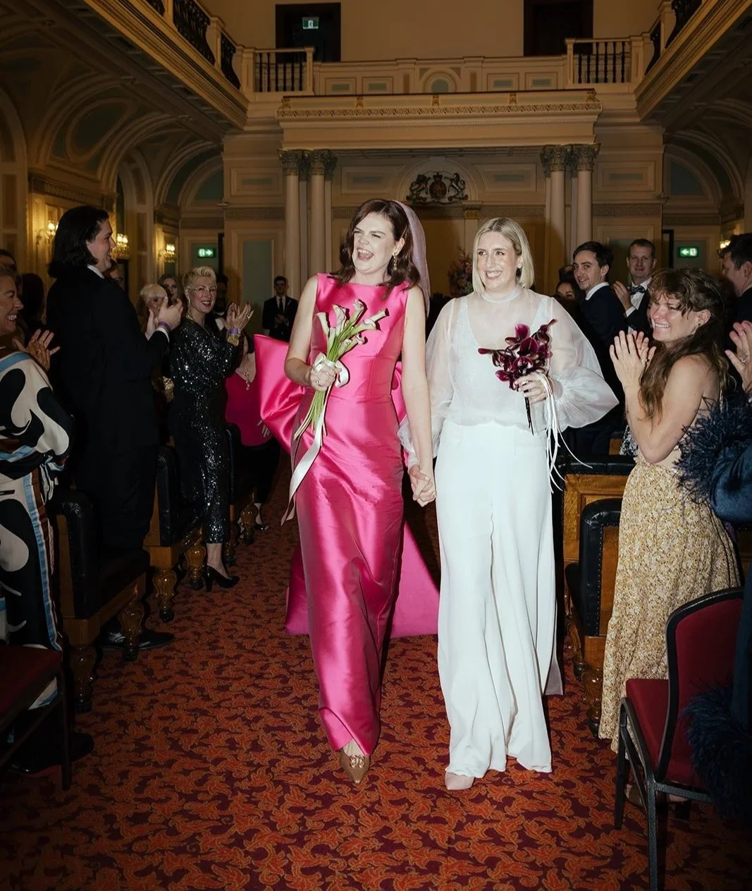 Two women in elegant dresses walking hand-in-hand through a celebration, surrounded by smiling and clapping people in a grand, ornate hall.