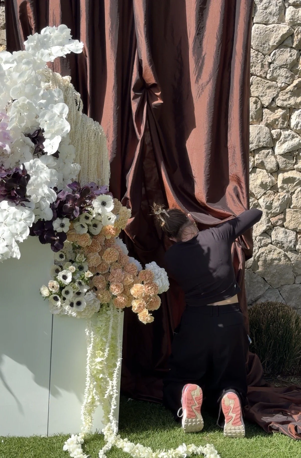 A woman kneeling on grass, arranging flowers and fabric for a decorative display near a stone wall and maroon curtain.