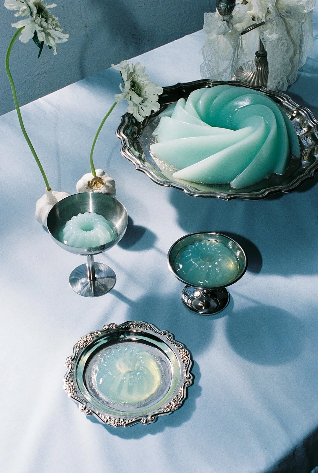 A table set with vintage silver dishes and two tall vases with white flowers, featuring mint green gelatin desserts in decorative molds.