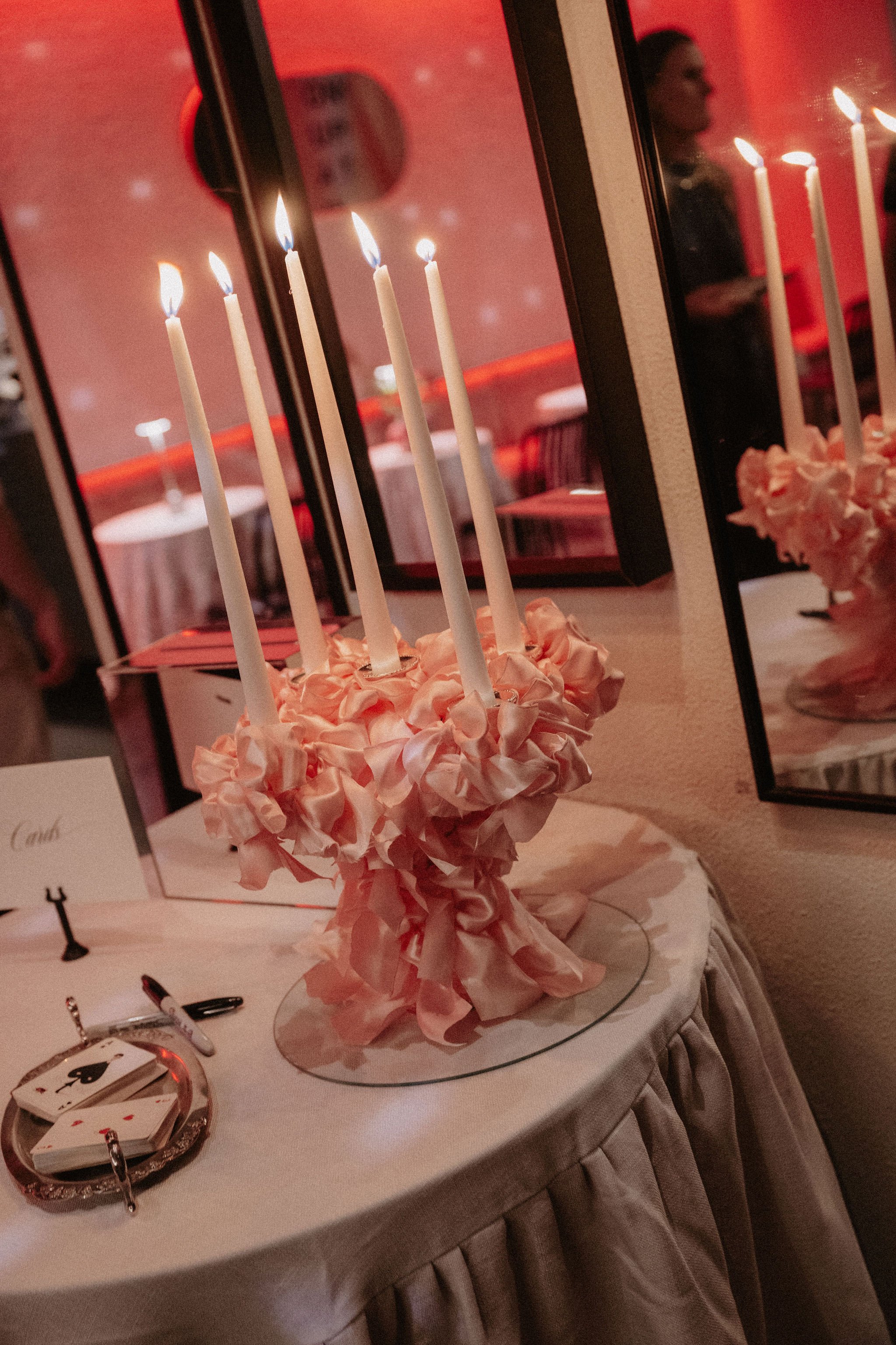 A table decorated with a pink napkin flower arrangement holding four lit white candles, with a mirror reflecting the candles and a woman in the background in a dimly lit room with pink lighting.