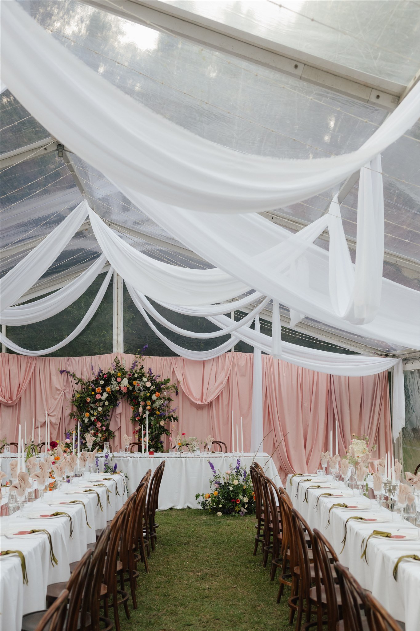 A decorated reception area with long tables covered in white tablecloths, set with plates and napkins, arranged in a U-shape on a grassy area. Wooden chairs surround the tables. A backdrop of pink drapes and floral arrangements is at the front. The ceiling is decorated with white fabric drapes hanging from above.