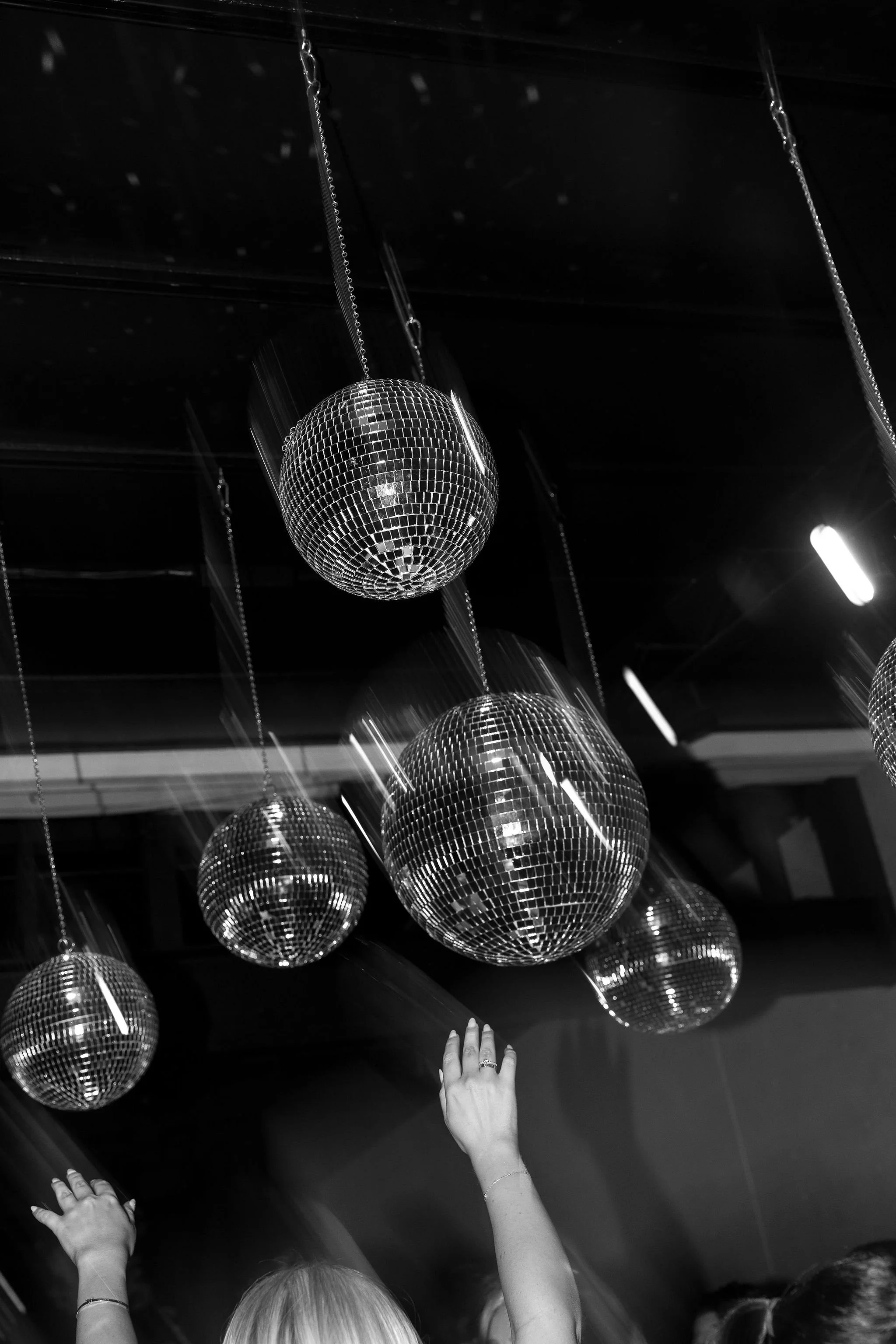 People at a party reaching towards hanging disco balls on the ceiling, in black and white photograph.