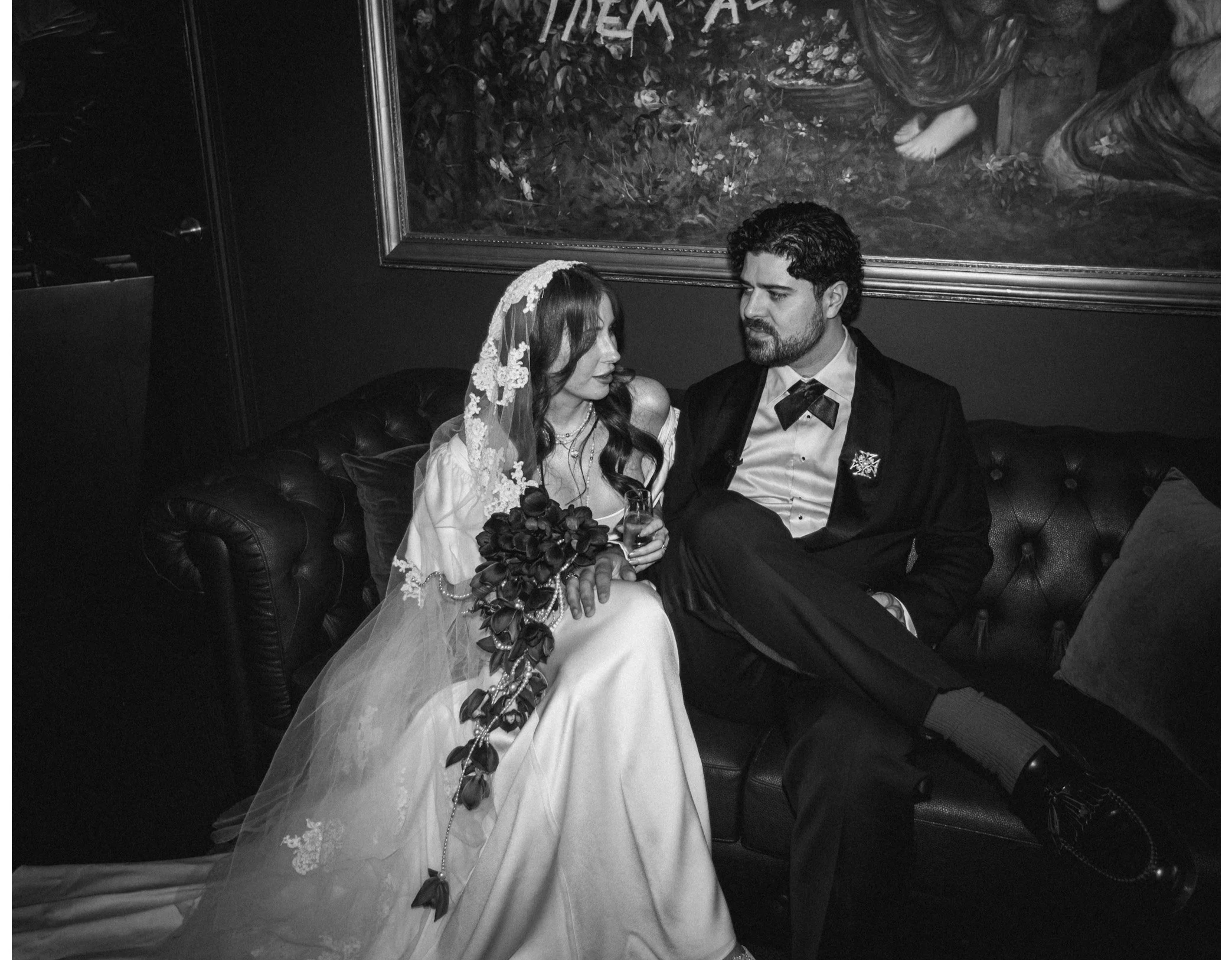A black and white photo of a couple seated on a leather sofa. The woman wears a wedding dress with lace and a veil, holding a bouquet. The man is dressed in a suit with a bow tie. Behind them is a framed painting on the wall and some plants.