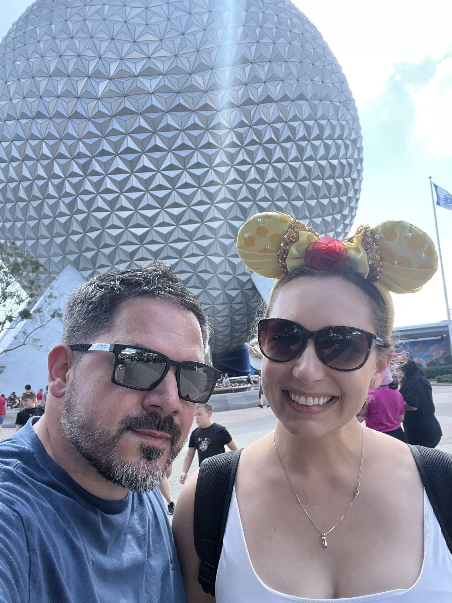 A man with glasses and a beard and a woman with sunglasses and a Minnie Mouse headband taking a selfie in front of Spaceship Earth at EPCOT.