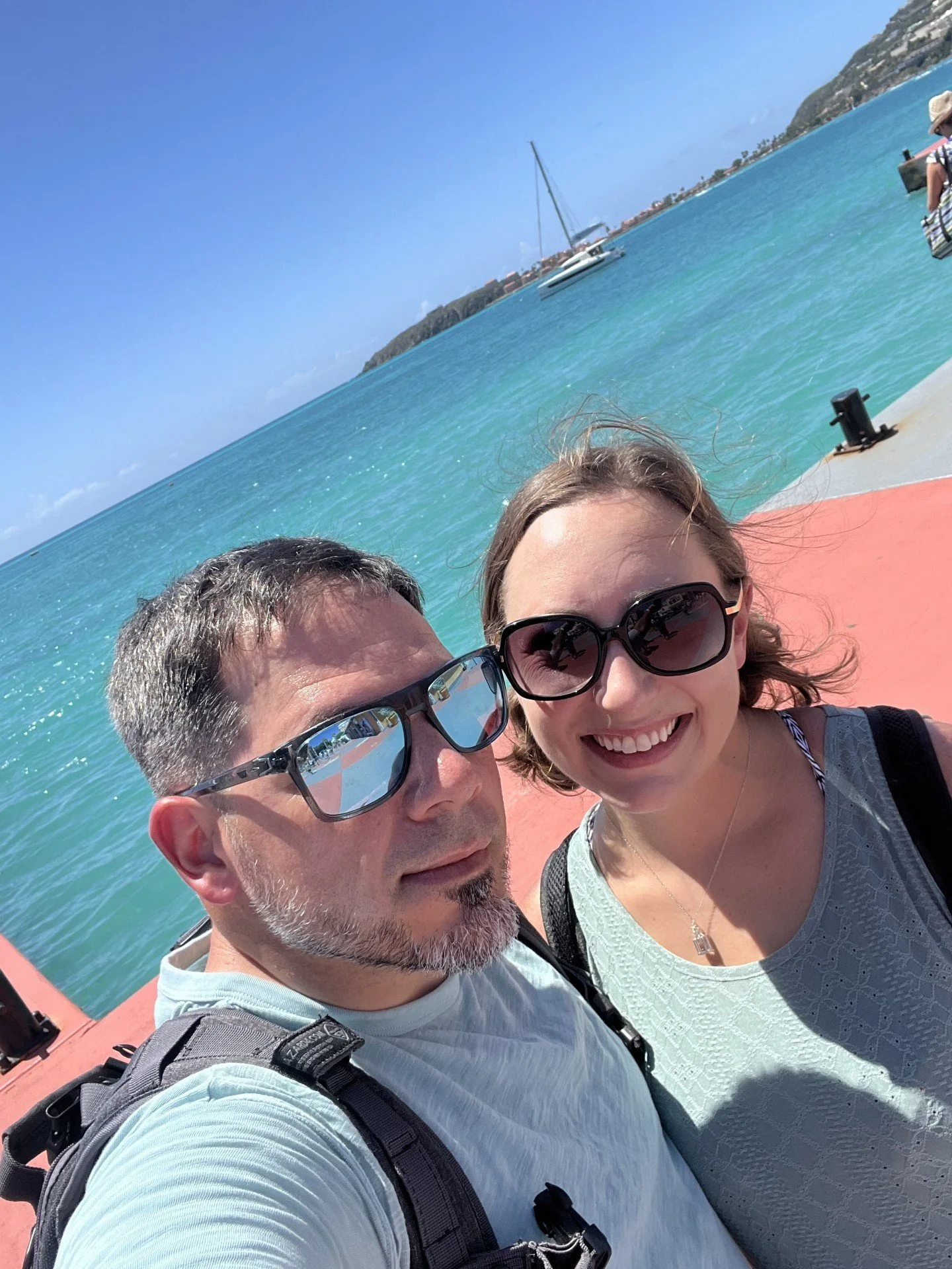 A smiling woman and man wearing sunglasses taking a selfie by the water, with a sailboat and distant shoreline in the background.