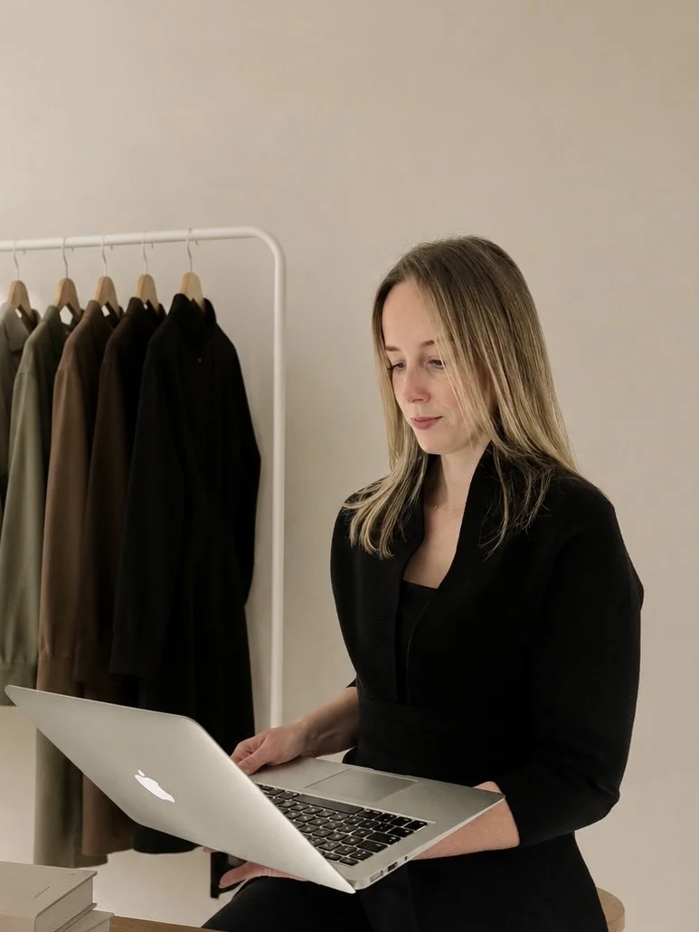 Fashion product development and sourcing for New Zealand fashion businesses. A woman in black clothing sitting and working on a silver MacBook laptop, with a clothing rack holding brown and black jackets in the background.