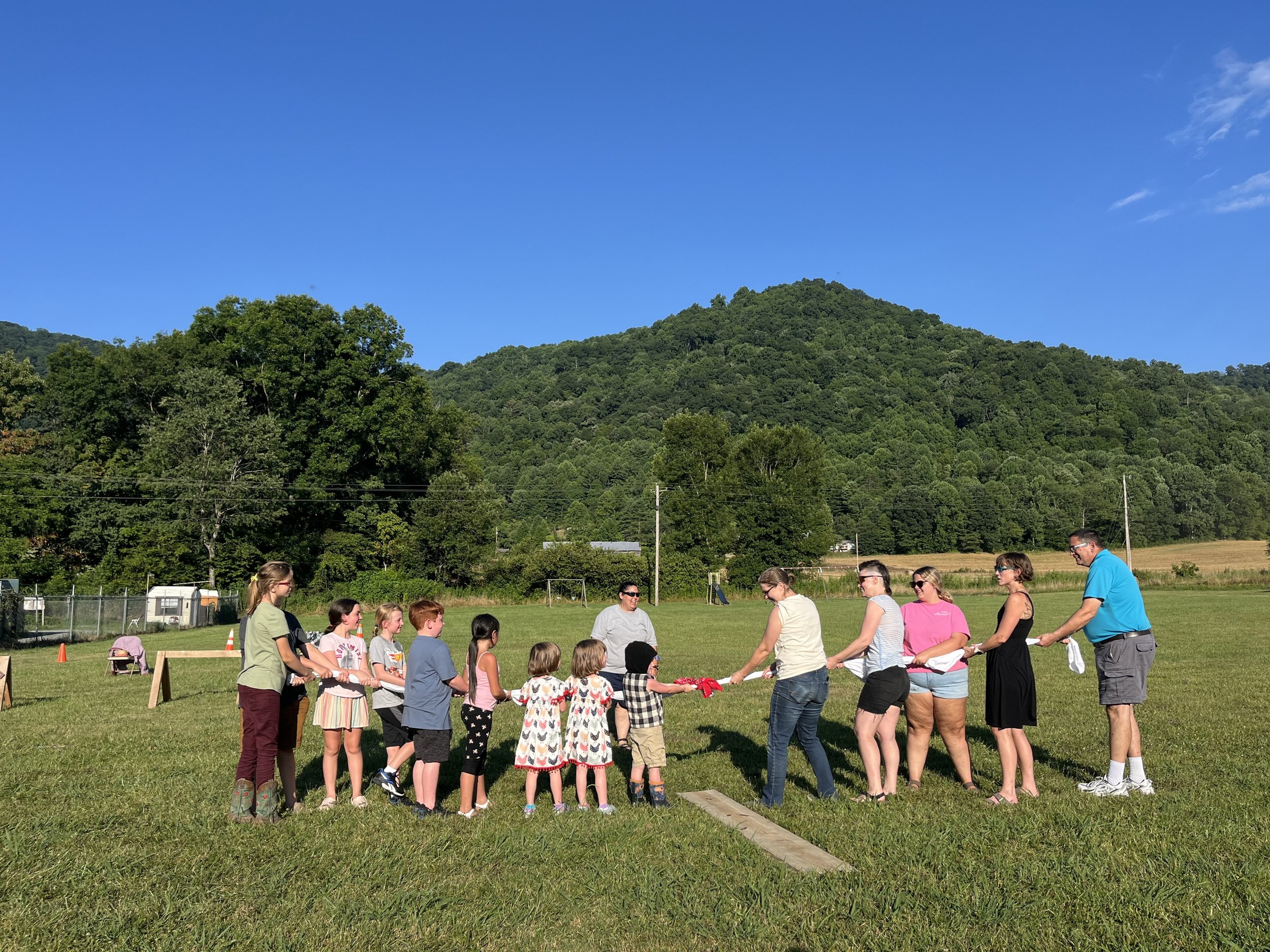 A group of children and adults standing in a line outdoors, holding a rope in a game of tug-of-war, on a grassy field with a backdrop of green trees and a mountain under a clear blue sky.