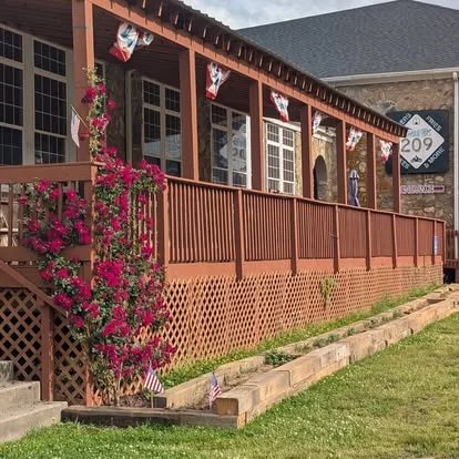 Front porch of a building decorated for the 4th of July with red, white, and blue banners, pink flowers, and small American flags in a flower bed.
