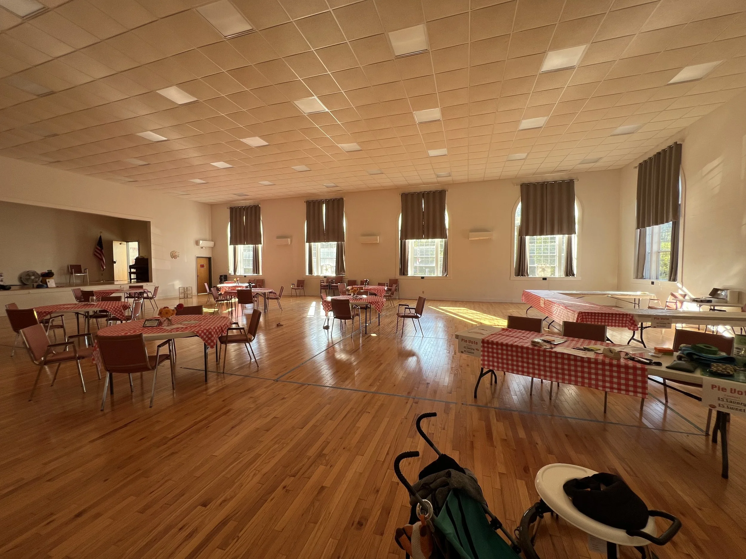 An indoor community hall or event space with a high ceiling and multiple large windows with curtains. The room has wooden floors and is set up with numerous tables covered with red and white checkered tablecloths, some with small floral centerpieces. There are chairs around the tables, and a few items and signs on some tables indicate that the space might be prepared for an event or gathering.
