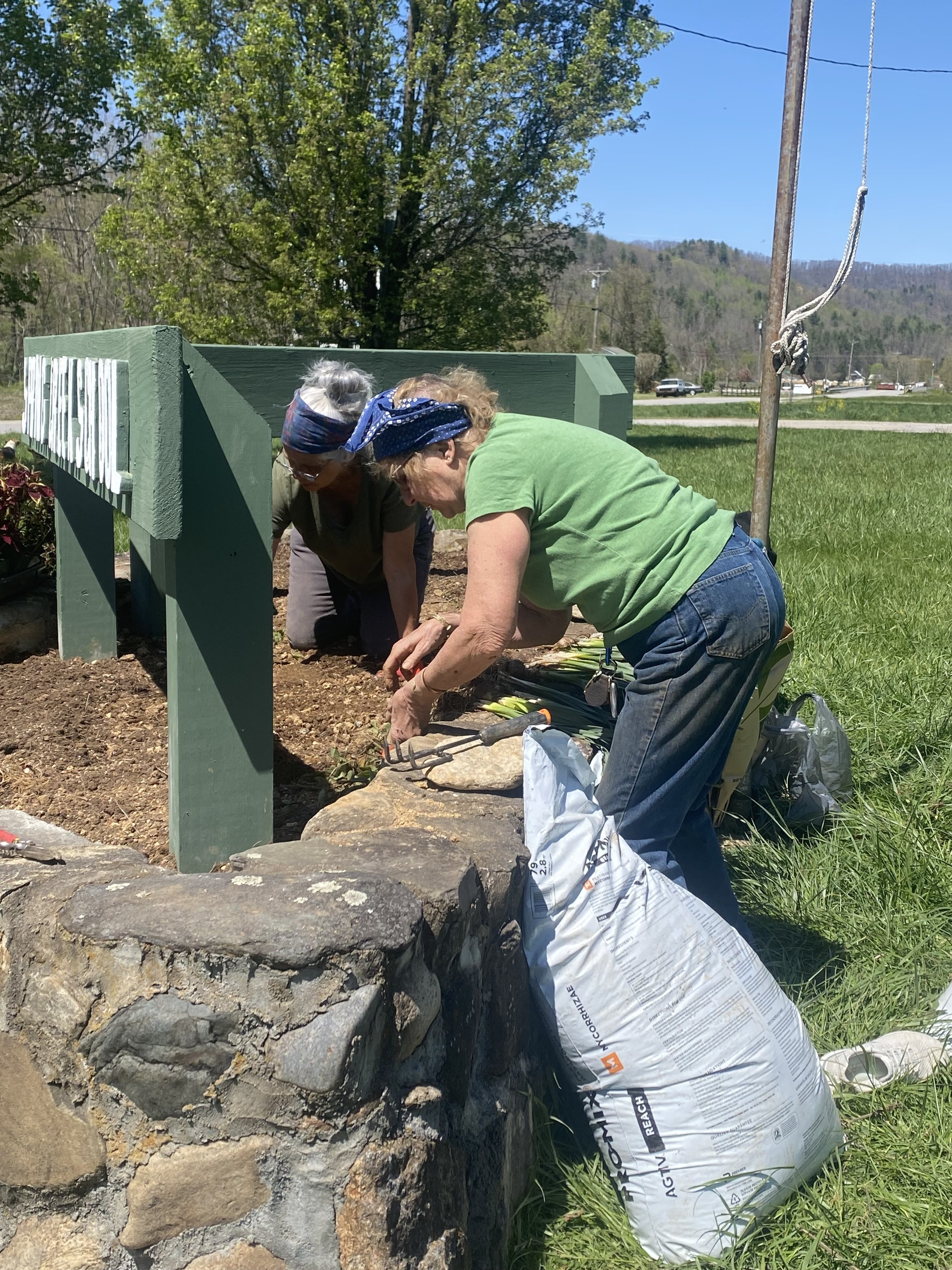 Two women working together planting flowers in the soil near a green wooden sign, with a rock border and a flag pole nearby, under a clear blue sky.