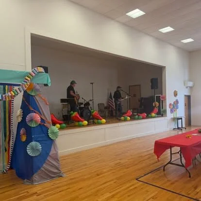 Decorated stage with balloons and fan decorations, with musicians preparing to perform and a red table with a red tablecloth in front of the stage.