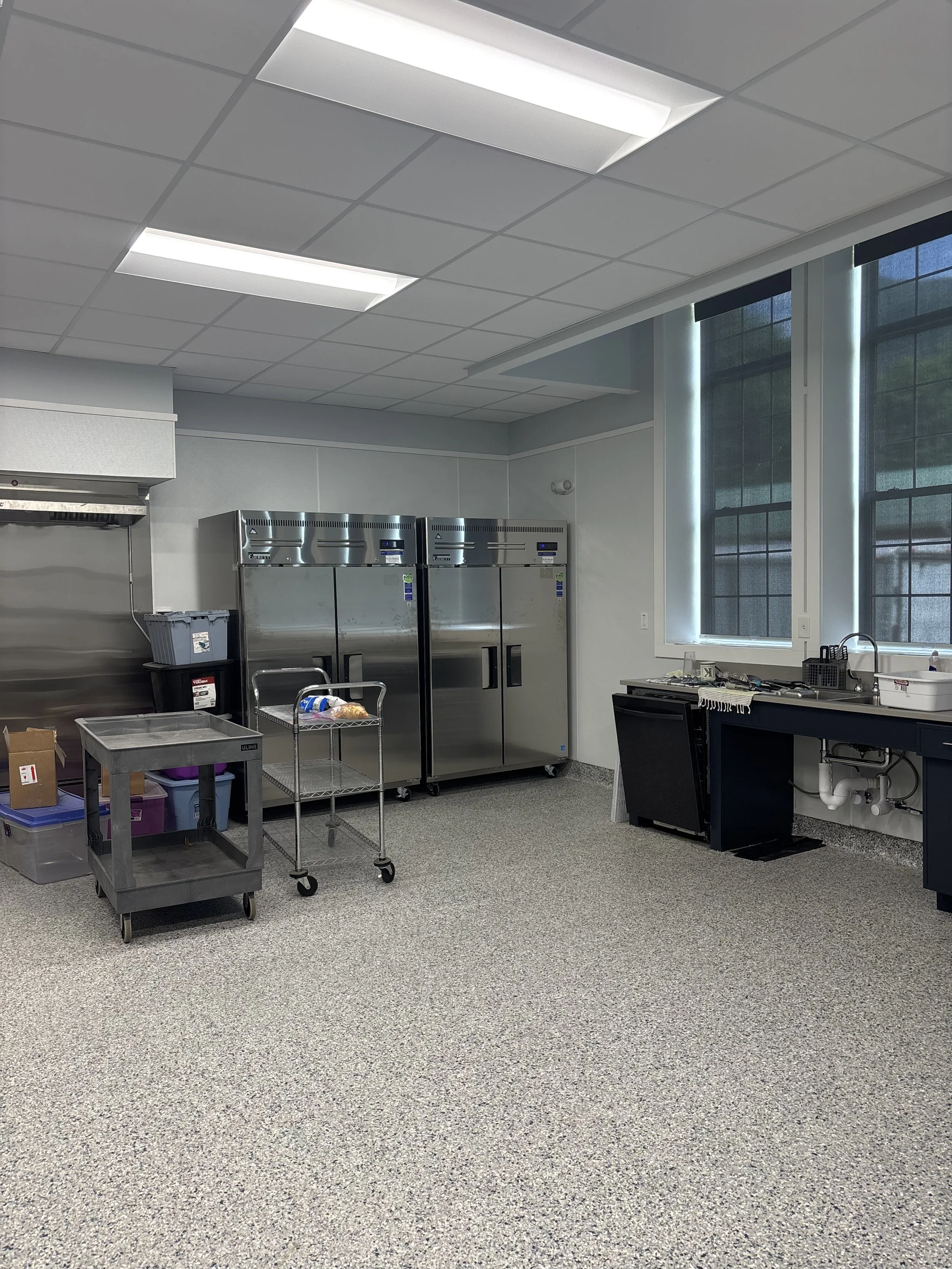 An empty commercial kitchen with stainless steel refrigerators, a black counter, a small table with supplies, large windows with blinds, and a gray speckled floor.