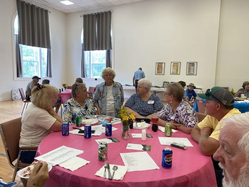 Group of elderly people and a man in a yellow shirt with a baseball cap sitting around a round table with a pink tablecloth, having a conversation during a social event in a bright room with large windows and curtains.