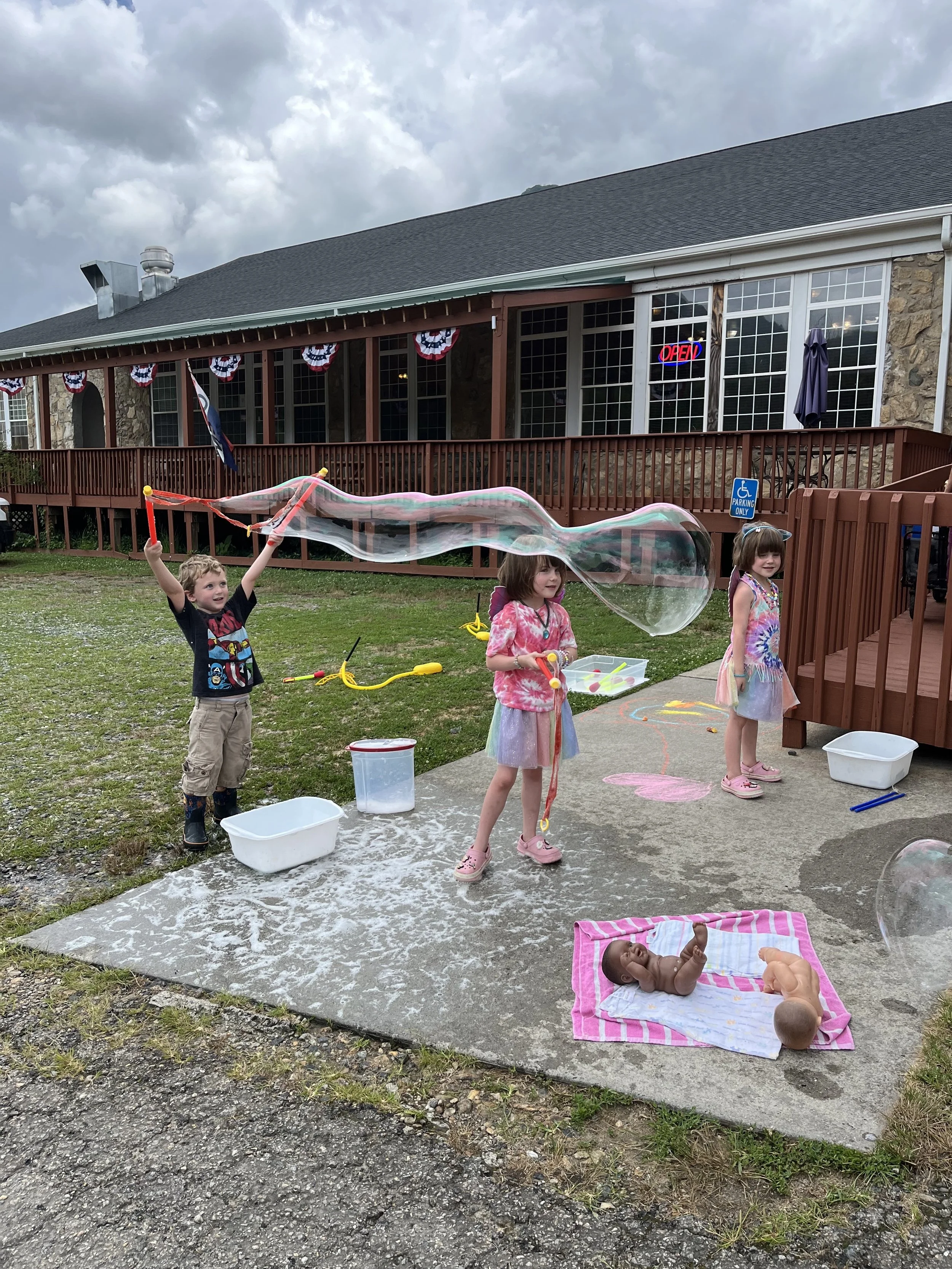 Children playing outside with soap bubbles, one child with arms raised holding a bubble wand, two children standing nearby, a baby lying on a pink and white striped towel, and a young child with glasses on a pink and rainbow tie-dye dress. A large house with a wooden balcony and an 'Open' sign is in the background.