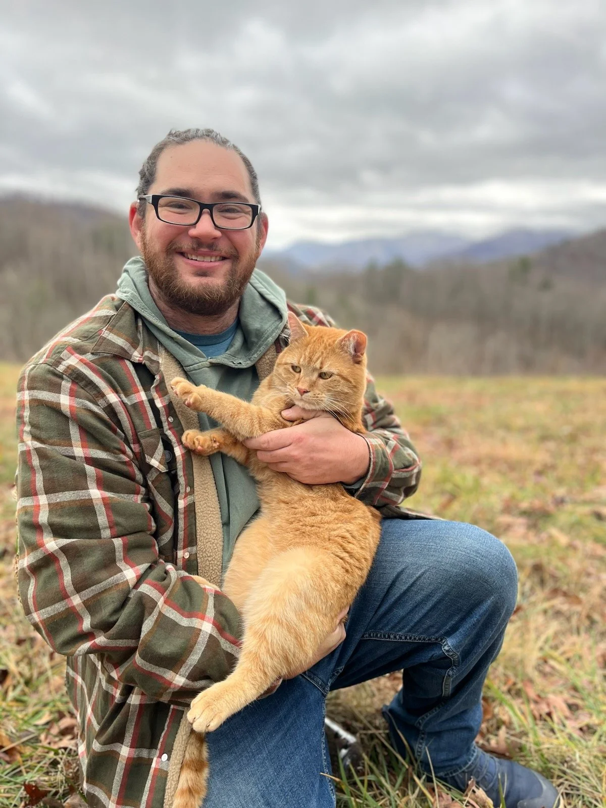 Man with glasses, beard, and long hair in a bun holding an orange tabby cat outdoors on a cloudy day.