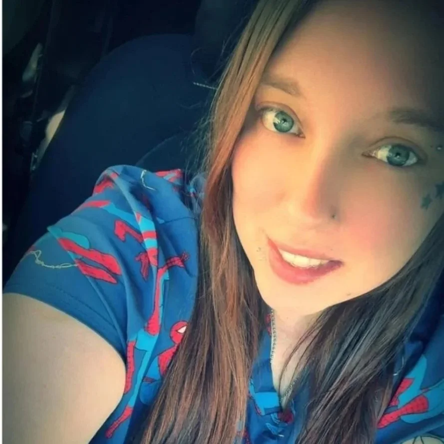 Close-up selfie of a woman with blue eyes and light brown hair, smiling and wearing a blue shirt with red and white patterns.