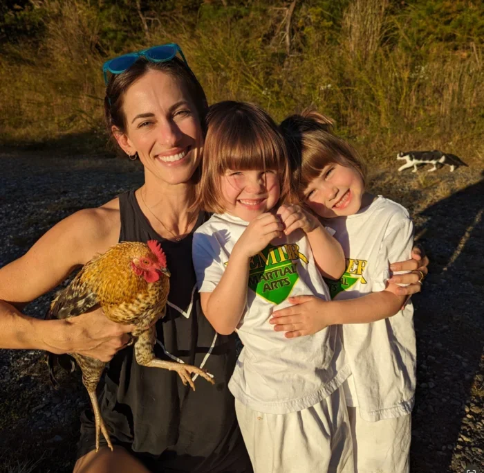 A woman and two young girls smiling and hugging outdoors, with the woman holding a chicken and a dog in the background.