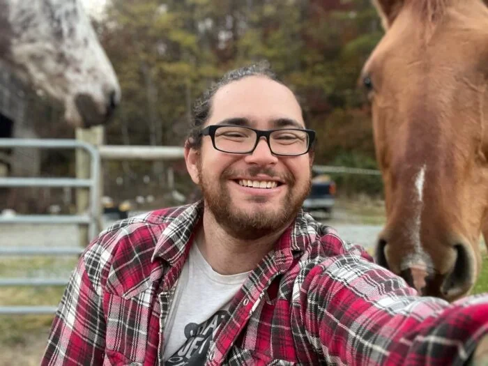 A smiling man with glasses and a beard taking a selfie with a horse on his right and a spotted horse in the background at a farm.
