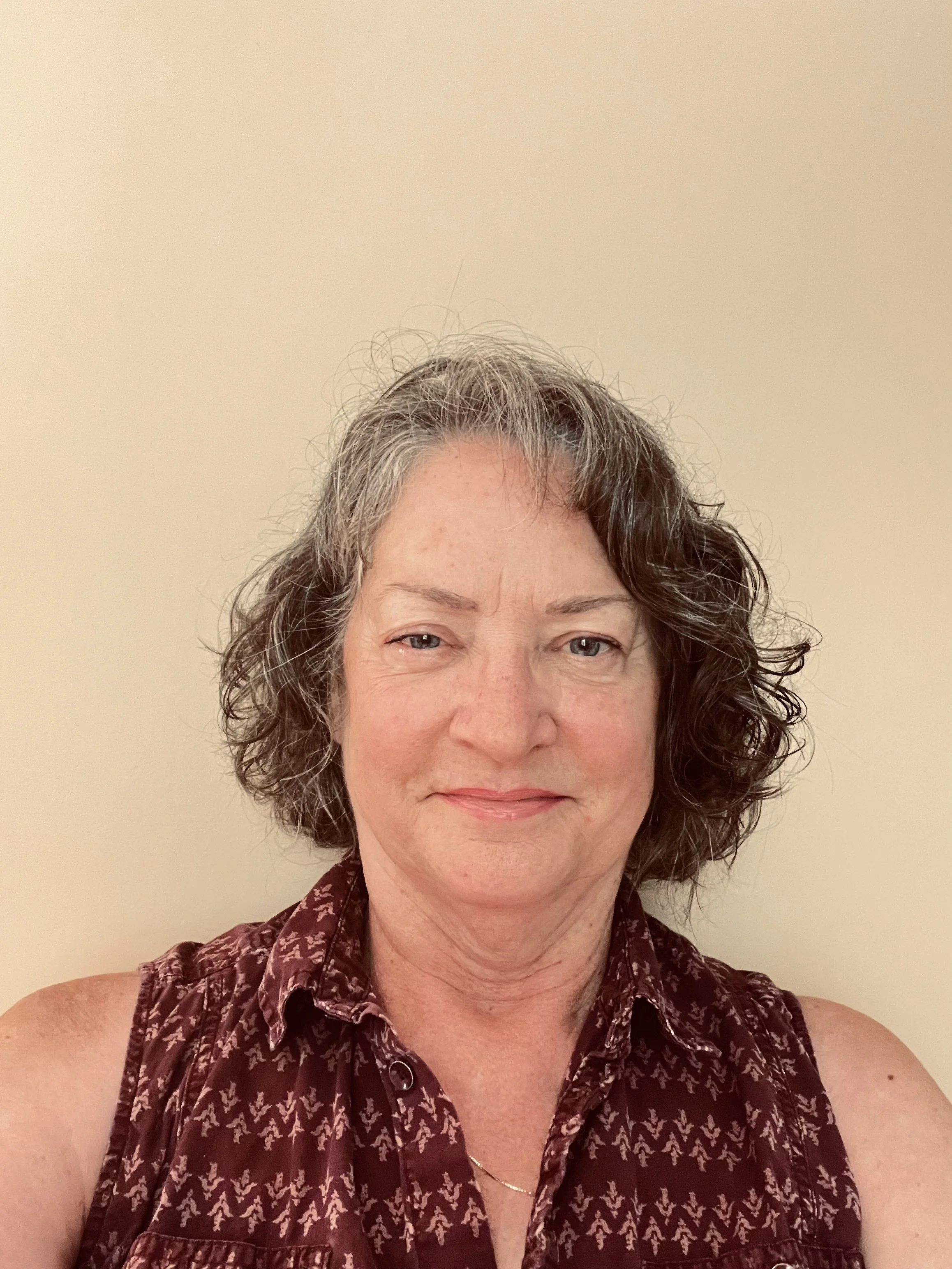 A middle-aged woman with wavy gray and brown hair, wearing a sleeveless maroon shirt with a pattern, standing against a plain beige wall.