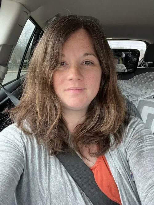 A woman with shoulder-length brown hair and fair skin taking a selfie inside a car, wearing a gray jacket over an orange shirt, with the seatbelt on. The background shows the car's interior and a rear window.