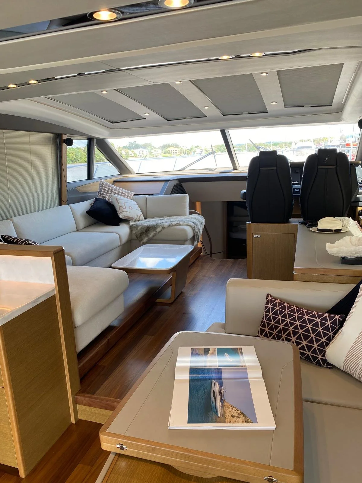 Interior of a yacht or boat with a beige sofa, pillows, a coffee table, another table with a magazine, two black captain chairs, and windows showing water and buildings outside.