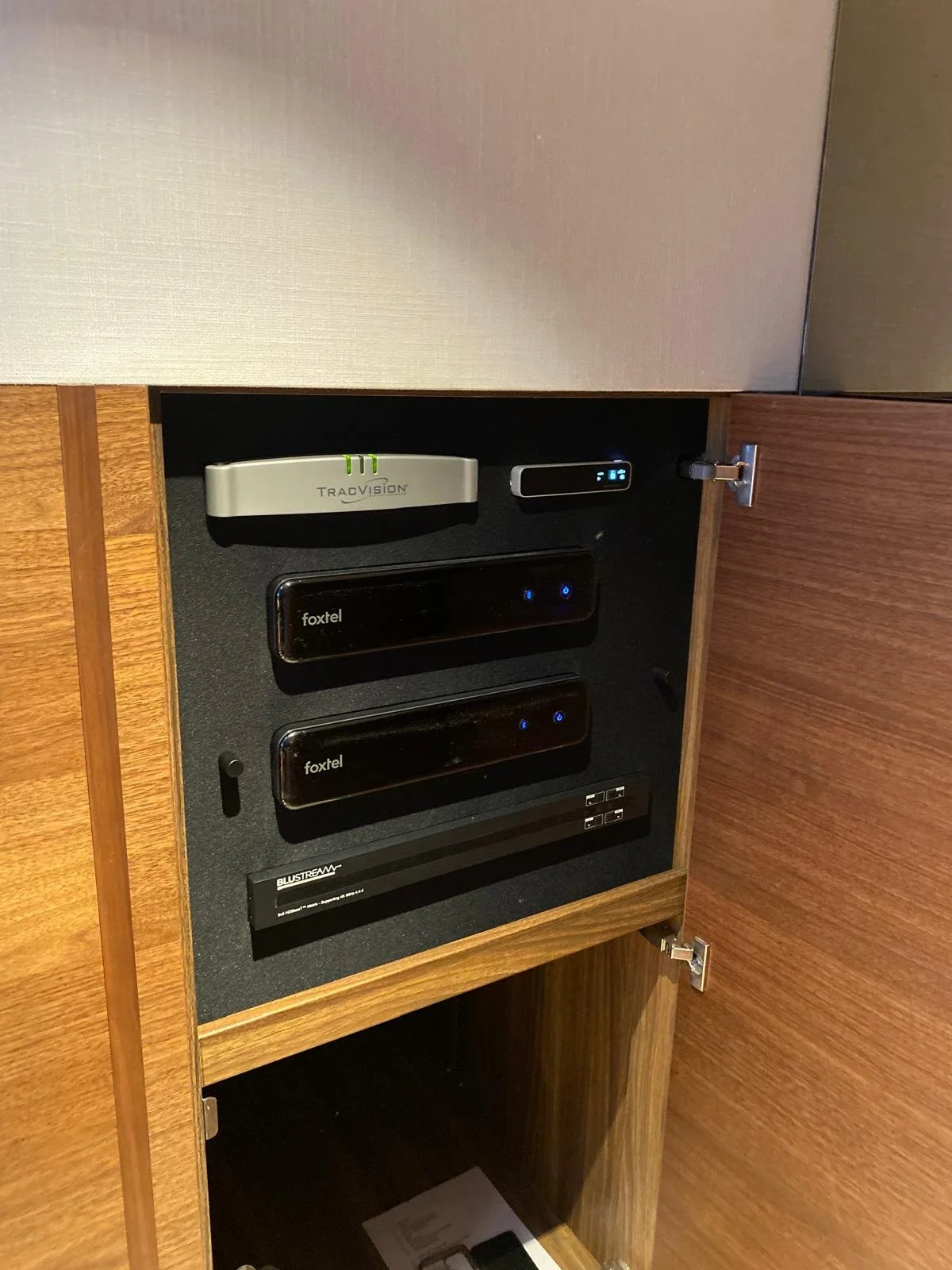 Open wooden cabinet containing electronic devices, including a TracVision satellite antenna controller, two Foxtel wireless boxes, and a Bluestream media player.