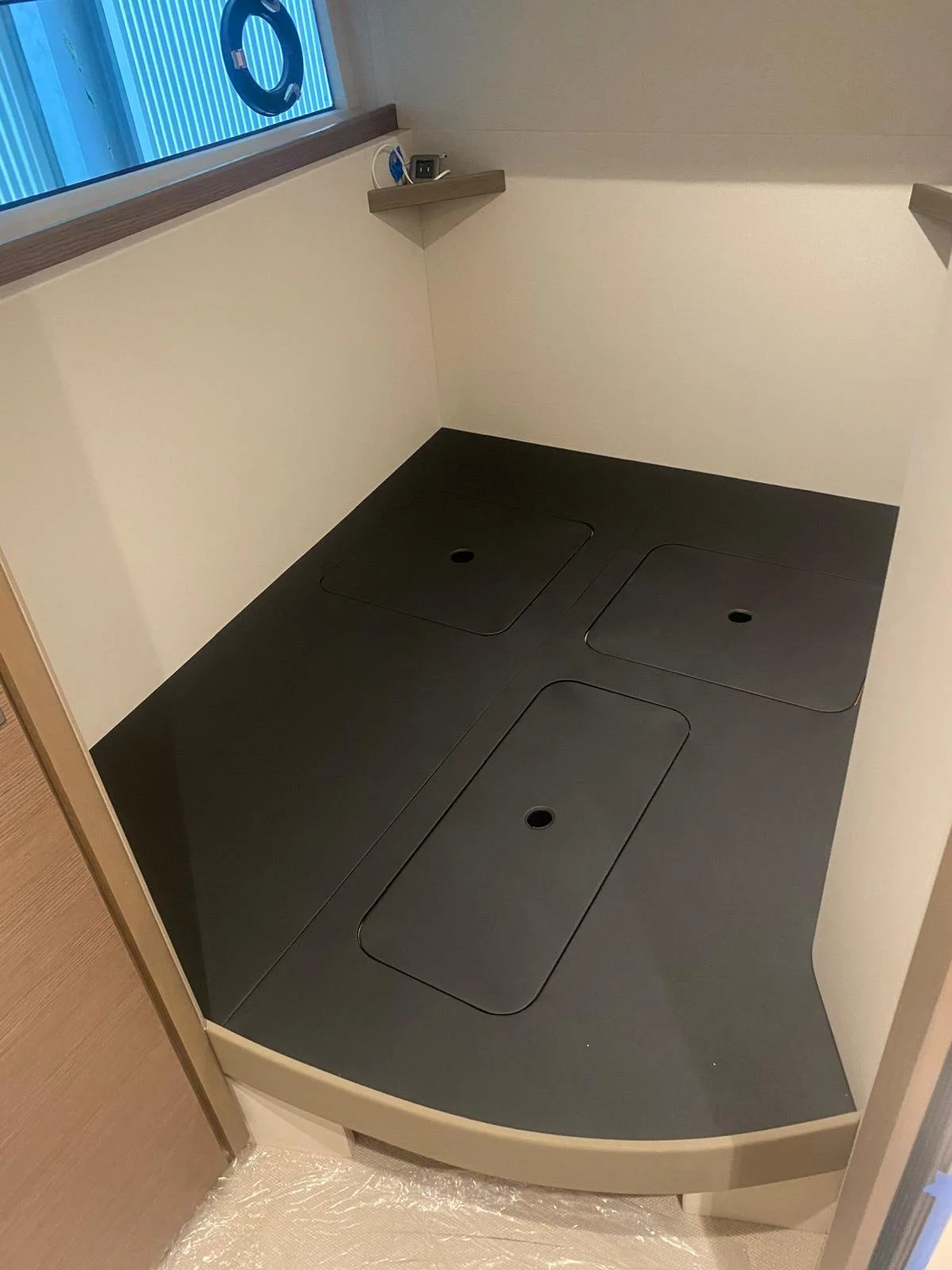 An empty shower with black access panels on the floor, a small shelf with an electrical outlet and plug, and a window with a blue towel hanging outside.