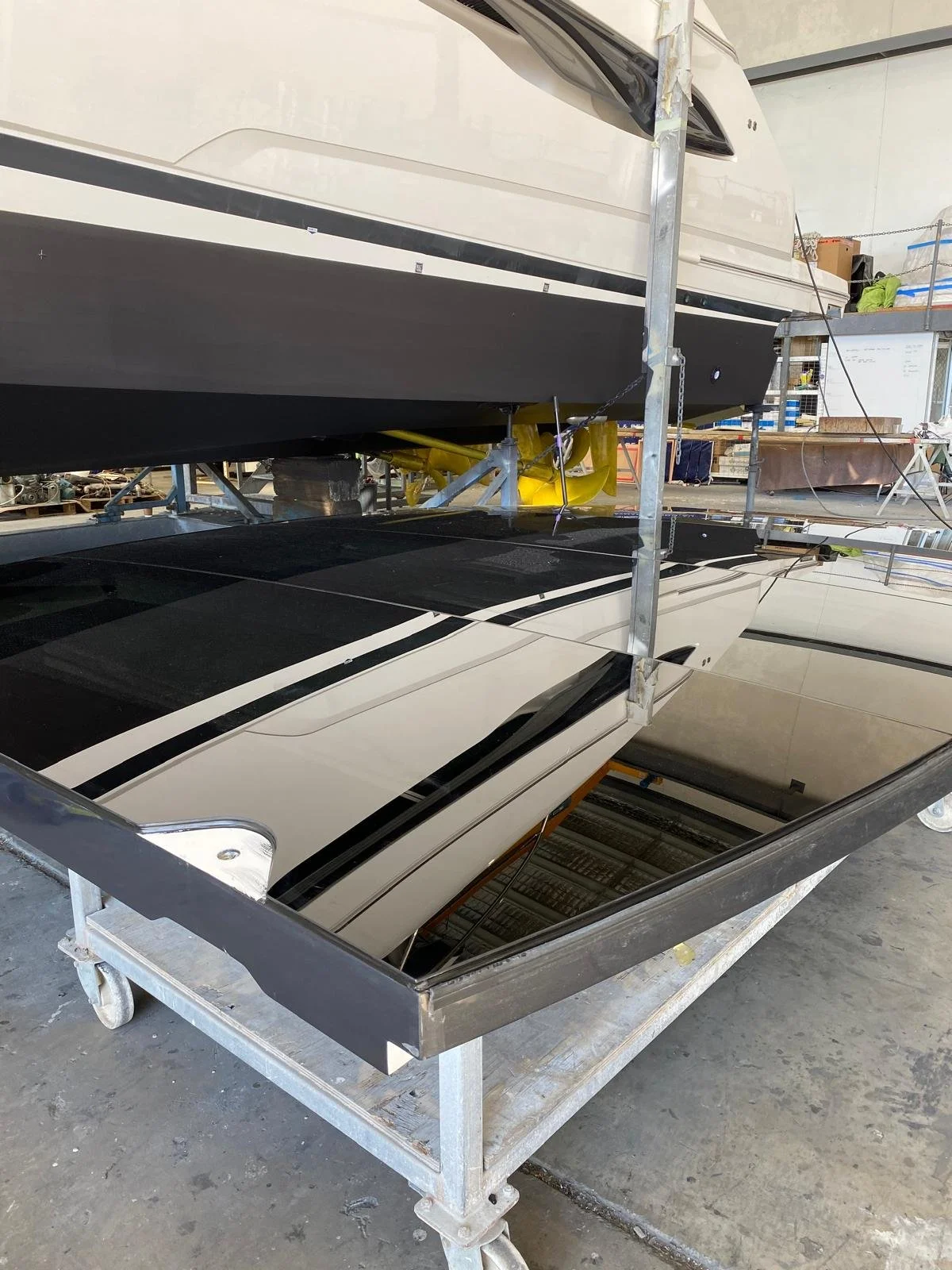 A boat inside a workshop, supported by scaffolding, with a black and white hull and a reflective mirrored surface beneath, showing parts of the workshop interior.