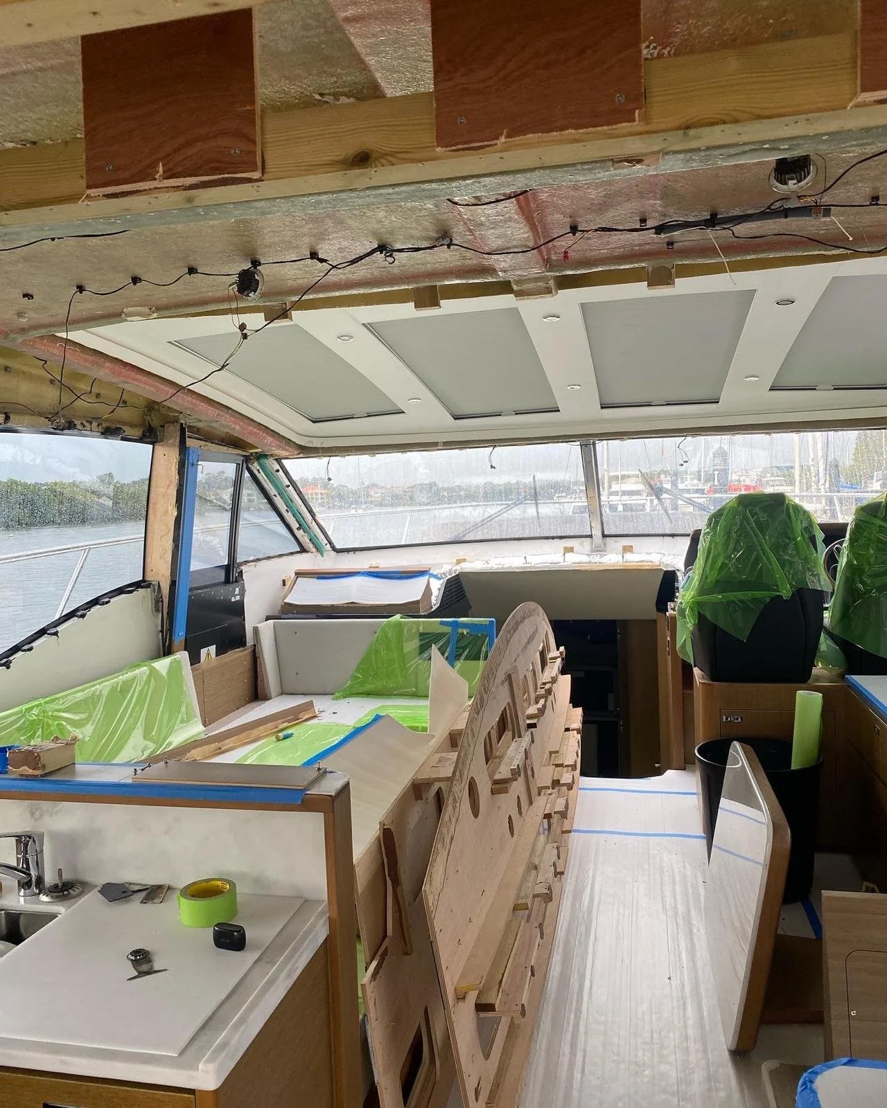Interior of a boat under construction with wooden framing, ceiling panels, and various tools and materials, including GREEN protective coverings on seats and construction supplies, with water visible outside the windows.
