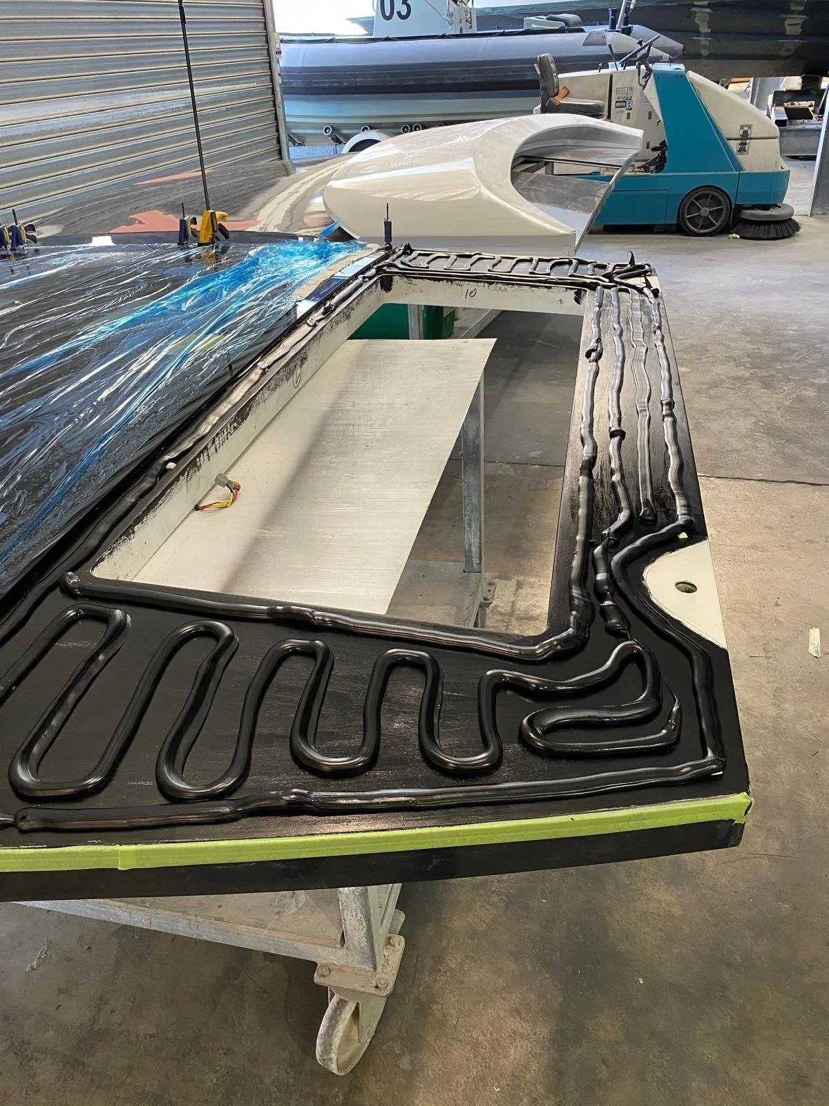 A work table with a metal frame, possibly the dashboard or part of a boat, featuring a black tubing pattern and electrical wiring. In the background, there is a white and blue boat, and a blue cleaning machine in a garage or workshop setting.