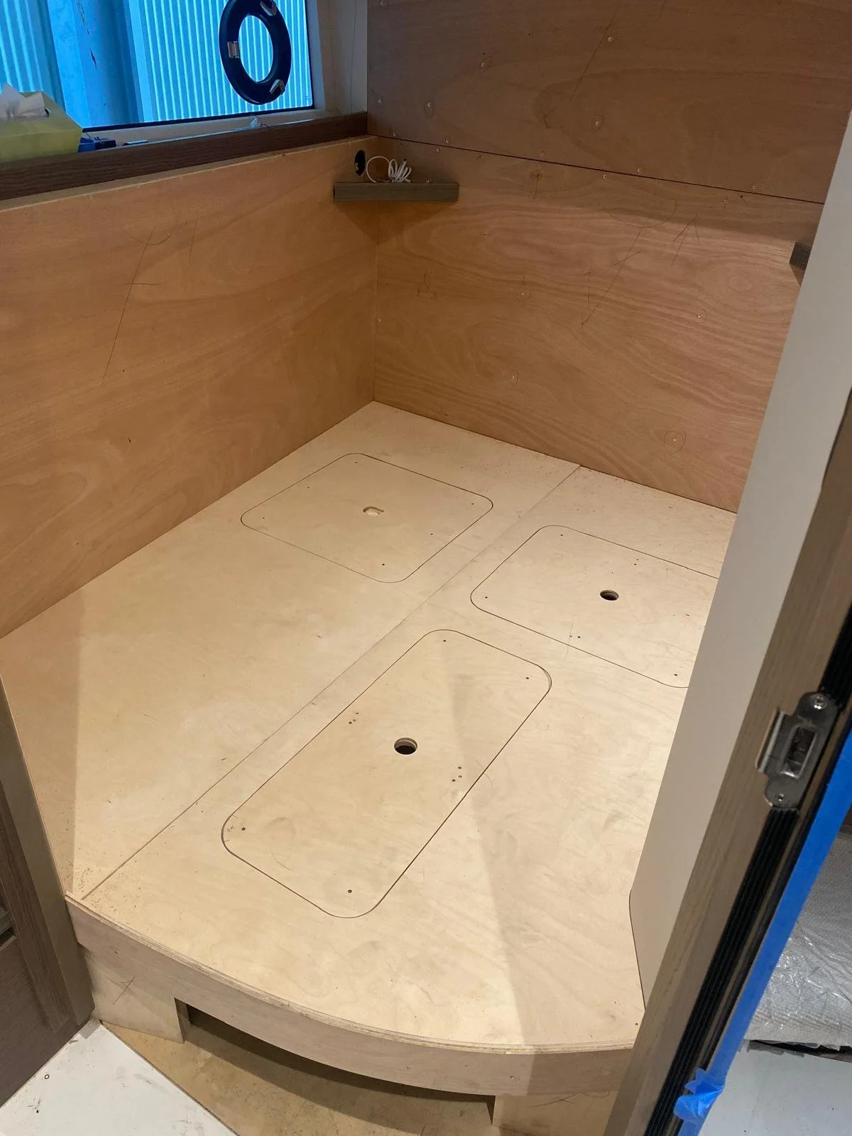 A small, empty, wooden shower with three access panels on the floor and a small shelf on the wall. There is a window with a blue blind above the shower.