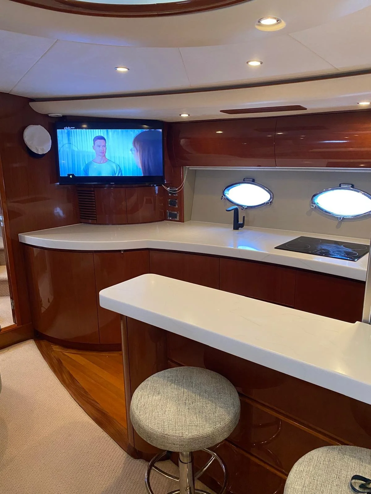 Interior of a yacht kitchen with wooden cabinets, a white countertop, a sink with a black faucet, a stove, a mounted flat-screen TV, and two round porthole windows.