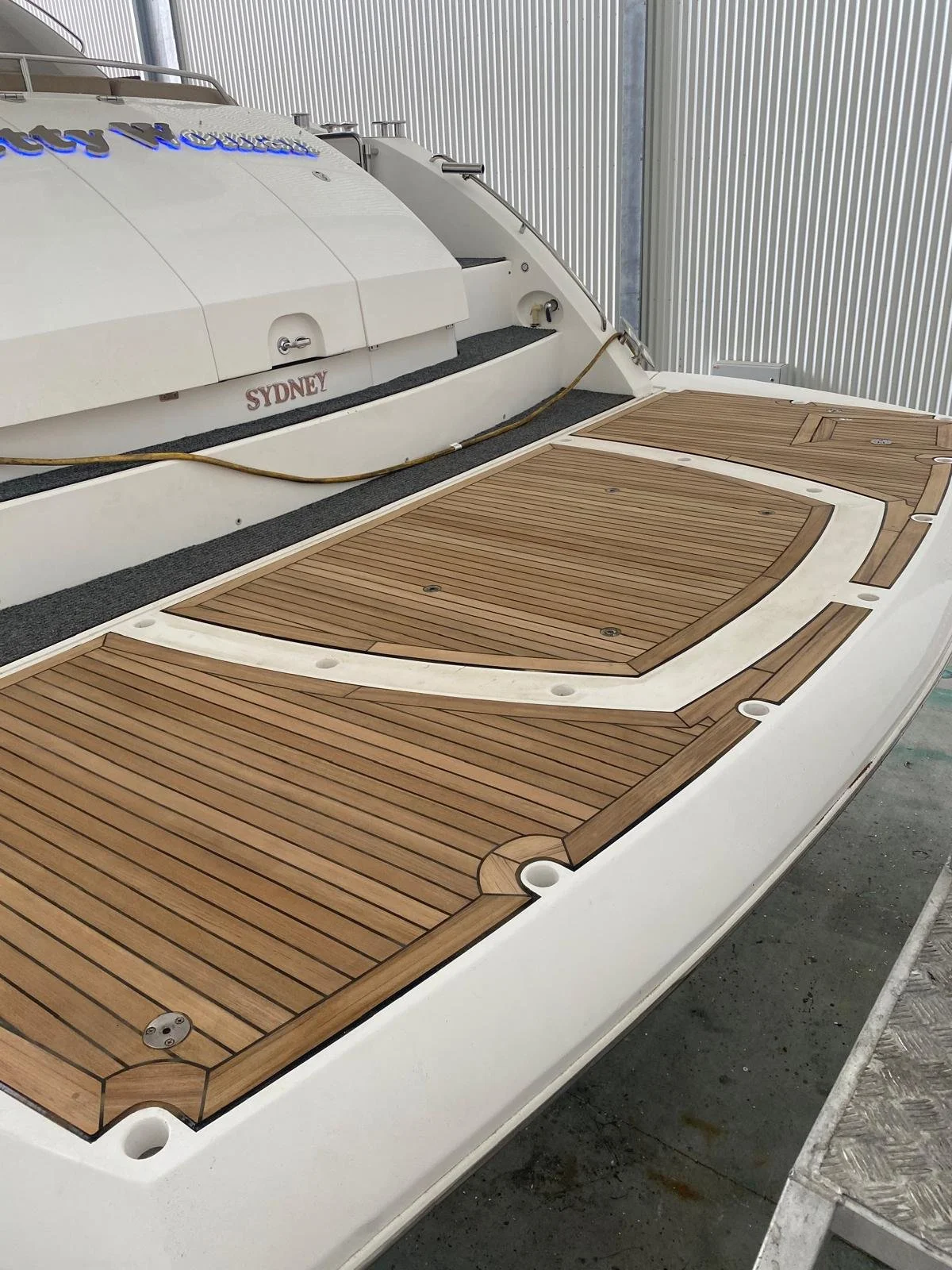 Close-up of the stern deck of a yacht with wooden and white non-slip surfaces, metal fasteners, and safety features, set against a corrugated metal wall.