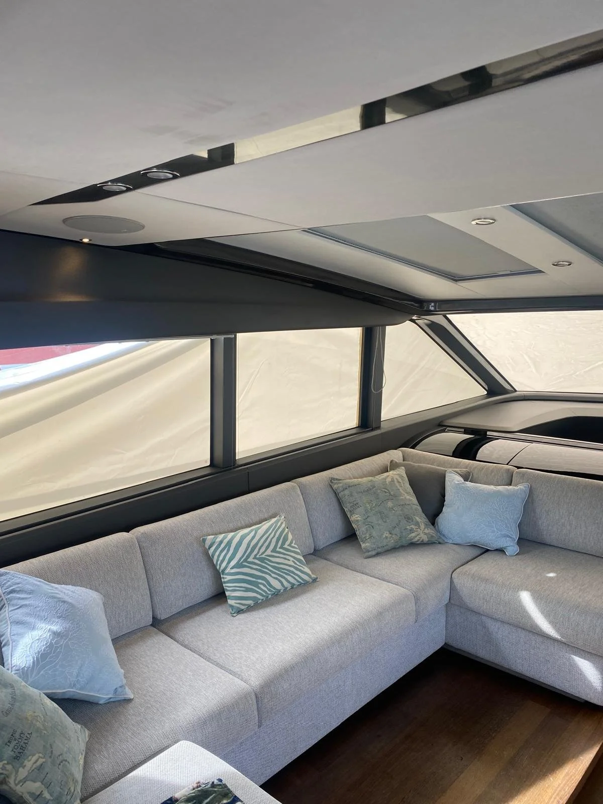 Interior of a yacht with a gray L-shaped sofa with patterned and solid pillows, large windows, and a ceiling with built-in lights.
