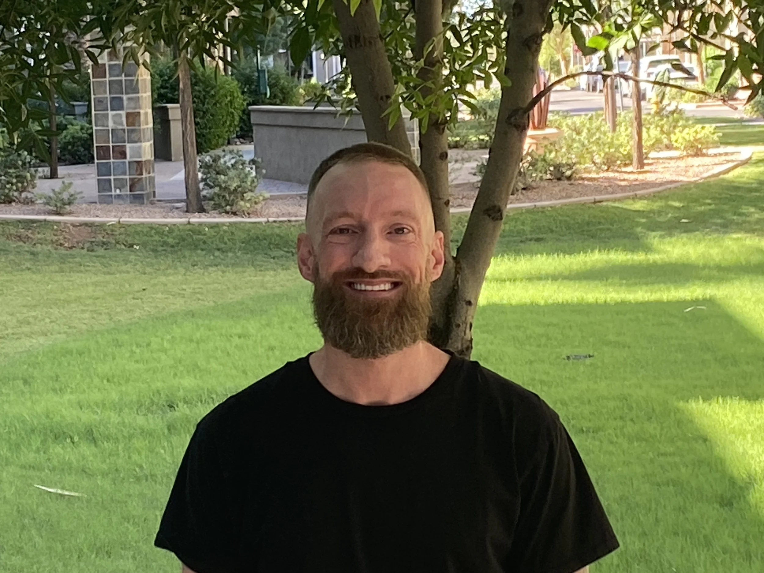 A man with a beard and smiling face standing outdoors in front of a tree, with greenery and a park-like setting in the background.