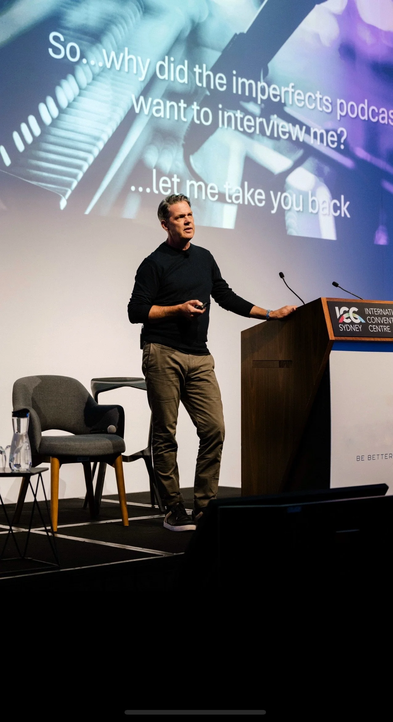A man standing on stage at the International Convention Centre in Sydney, giving a presentation. Behind him is a large screen displaying a humorous message about podcast interviews and a close-up of piano keys. There are chairs and a water pitcher on the stage.