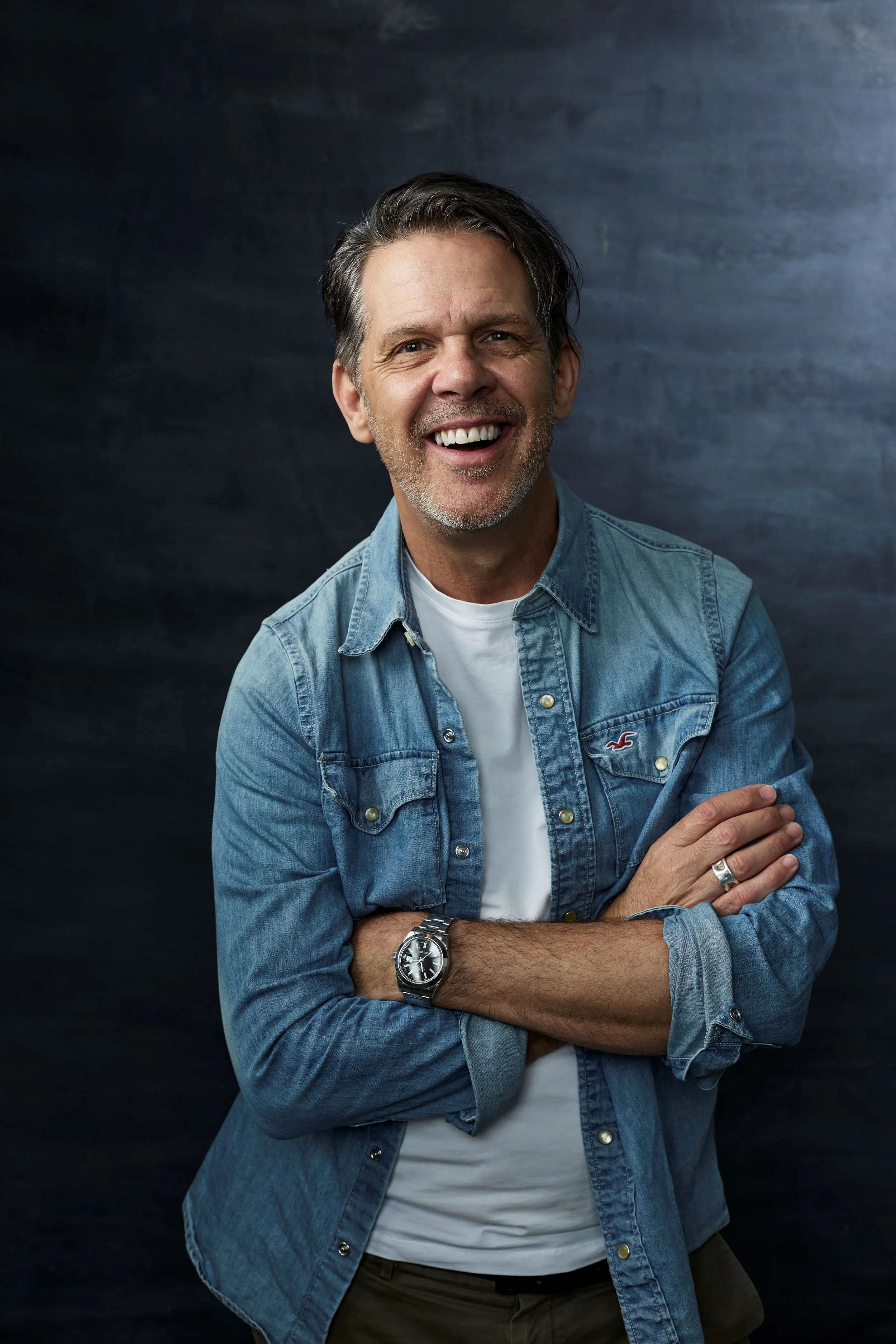 A joyful middle-aged man with gray hair, a stubble beard, wearing a denim shirt over a white t-shirt, smiling, with arms crossed, standing against a dark background.