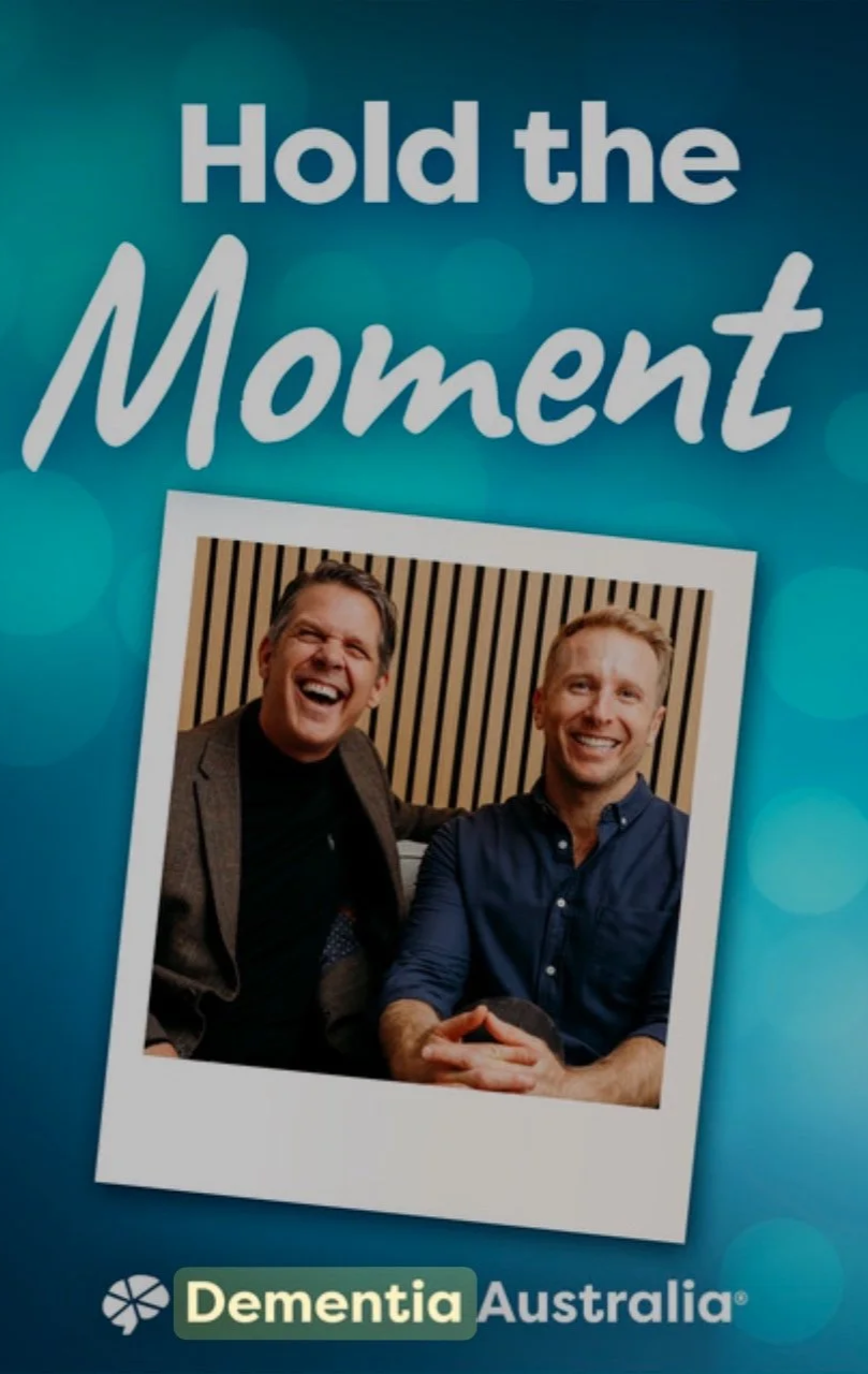 A promotional poster with the phrase 'Hold the Moment' at the top, a photograph of two men smiling and sitting together, and the logo for Dementia Australia at the bottom.
