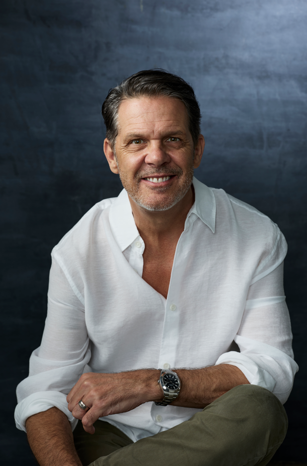 Portrait of a middle-aged man with dark hair and a beard, smiling, wearing a white button-up shirt with rolled-up sleeves and a watch, sitting against a dark blue textured background.